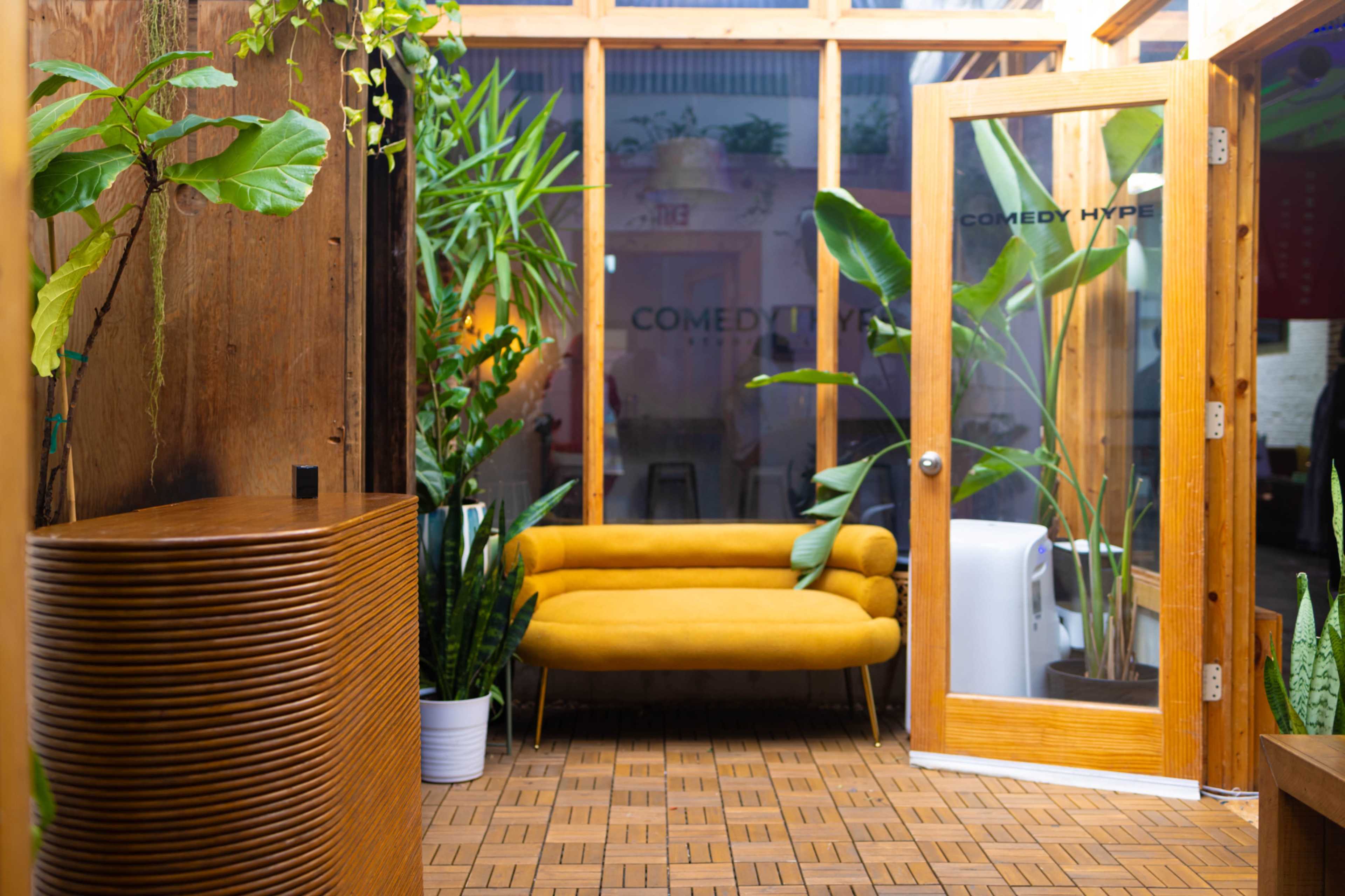 The image shows a bright indoor space with a wooden accent wall, a yellow couch, and various potted plants, with a glass door labeled "COMEDY HYPE."