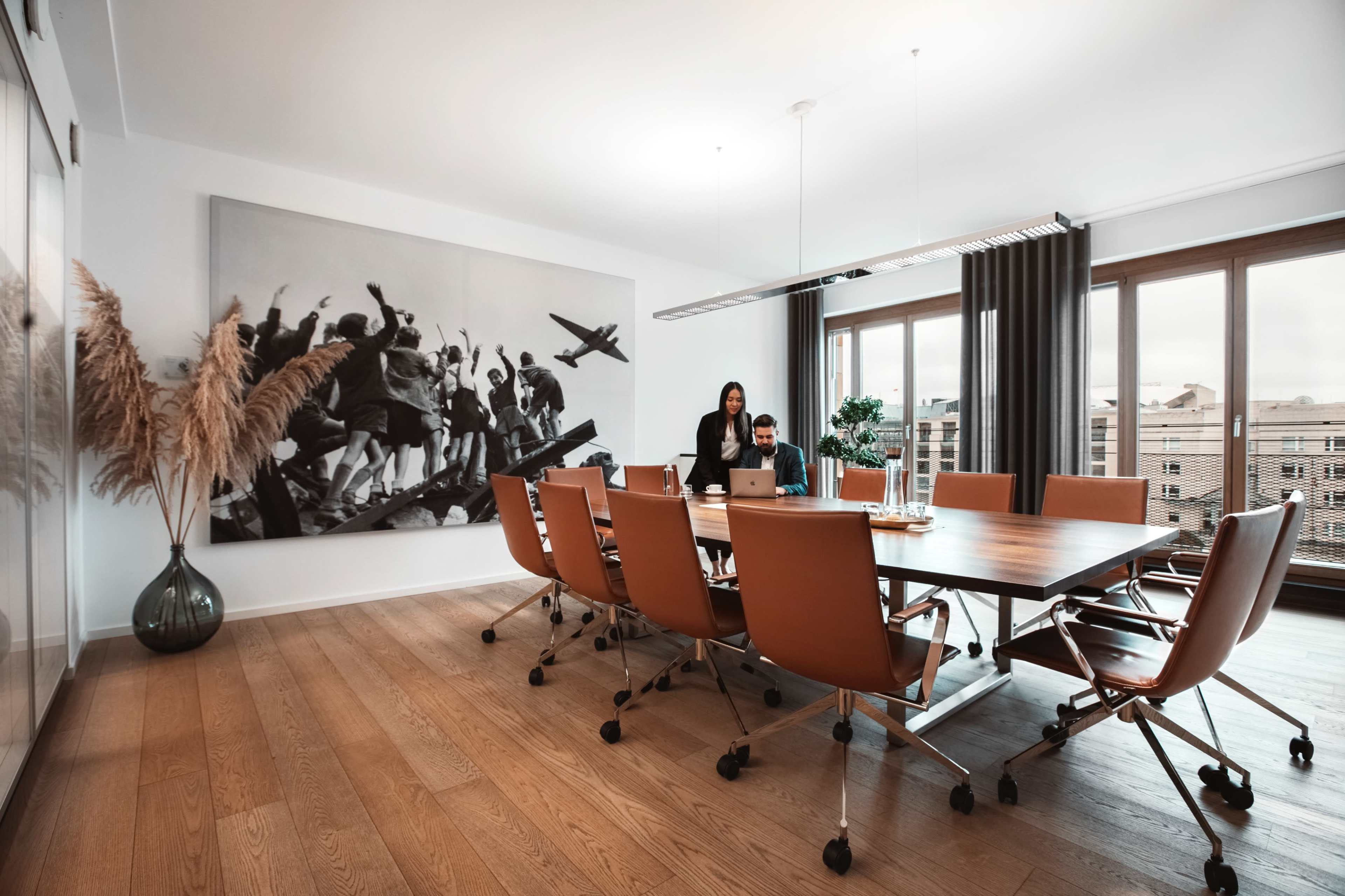 A modern conference room features a large wooden table surrounded by orange chairs, with a black-and-white mural on the wall and a group of people engaged in discussion.
