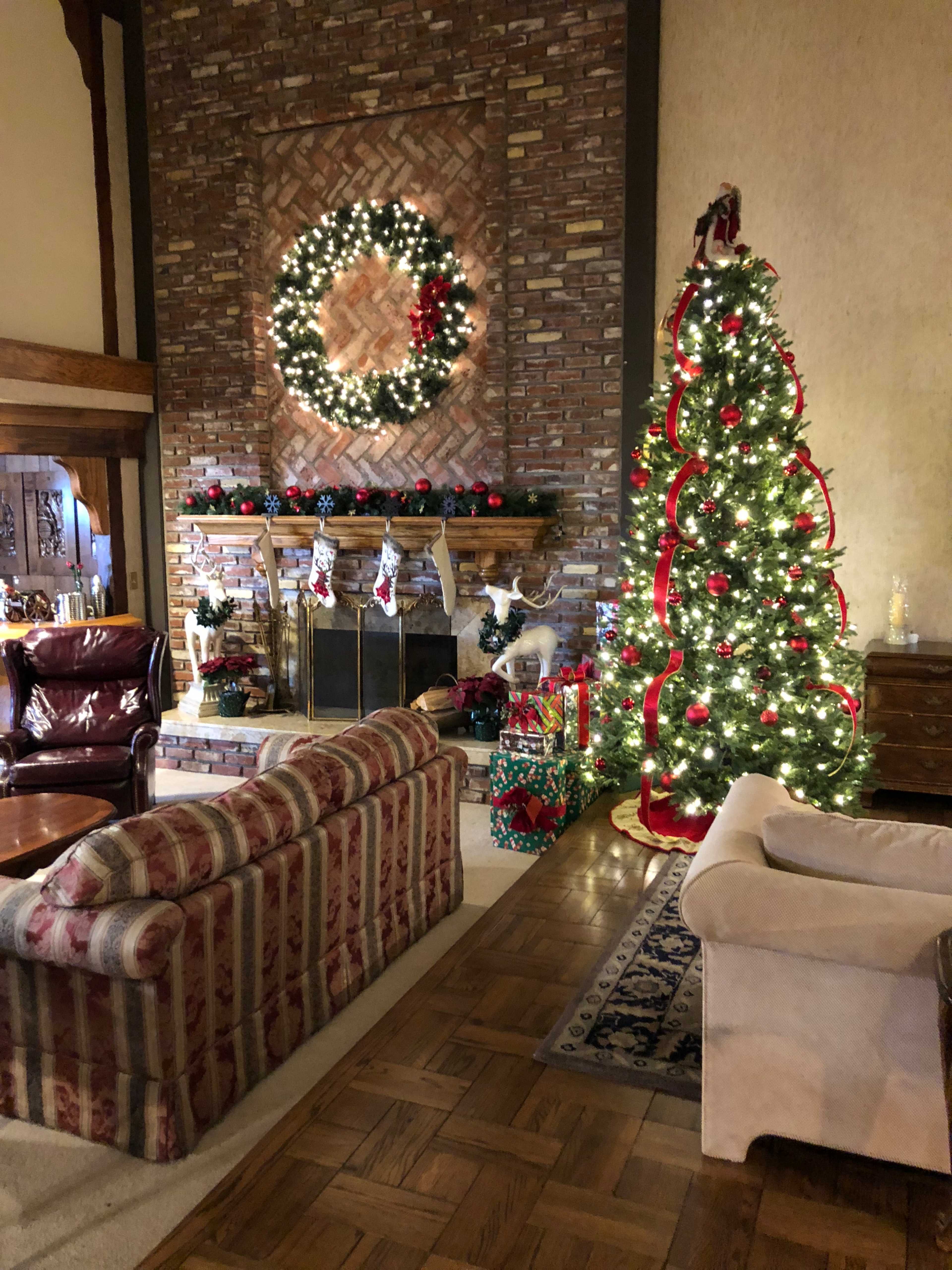 A warmly decorated living room features a stone fireplace adorned with a wreath and stockings, alongside a Christmas tree with lights and ornaments.
