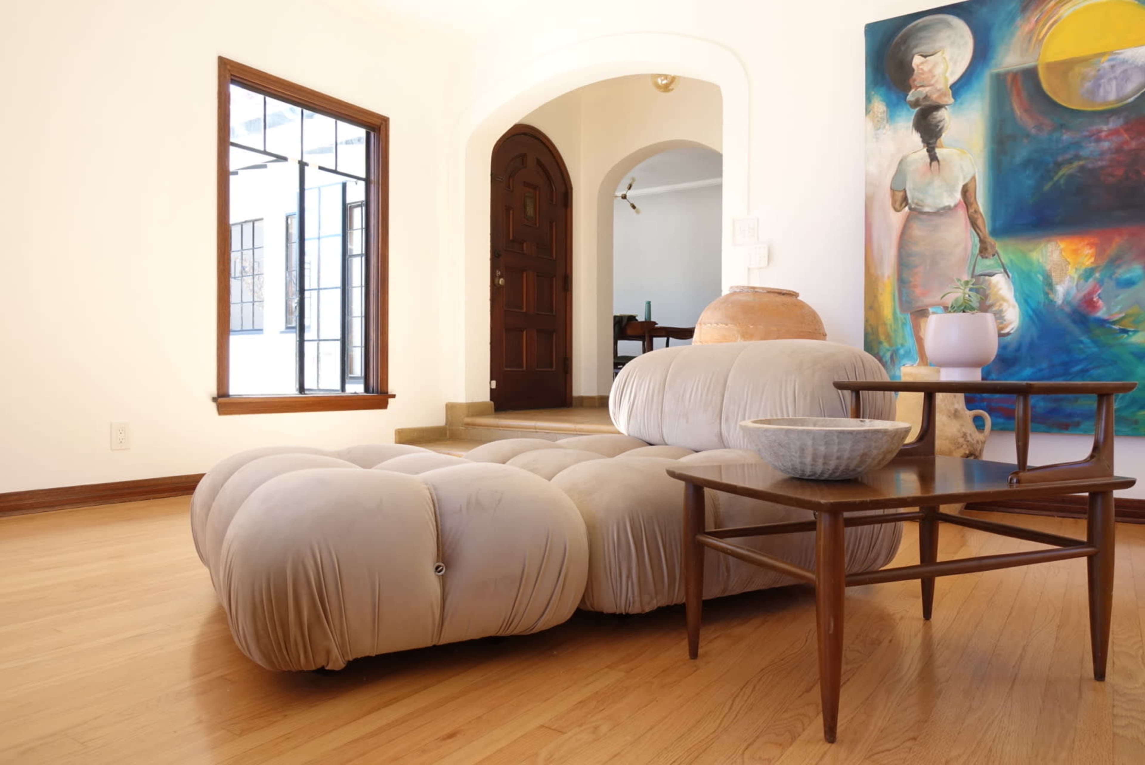 The image depicts a light-colored modern seating area with a plush sofa, a wooden table, and a vibrant painting on the wall, all set within a well-lit interior space.