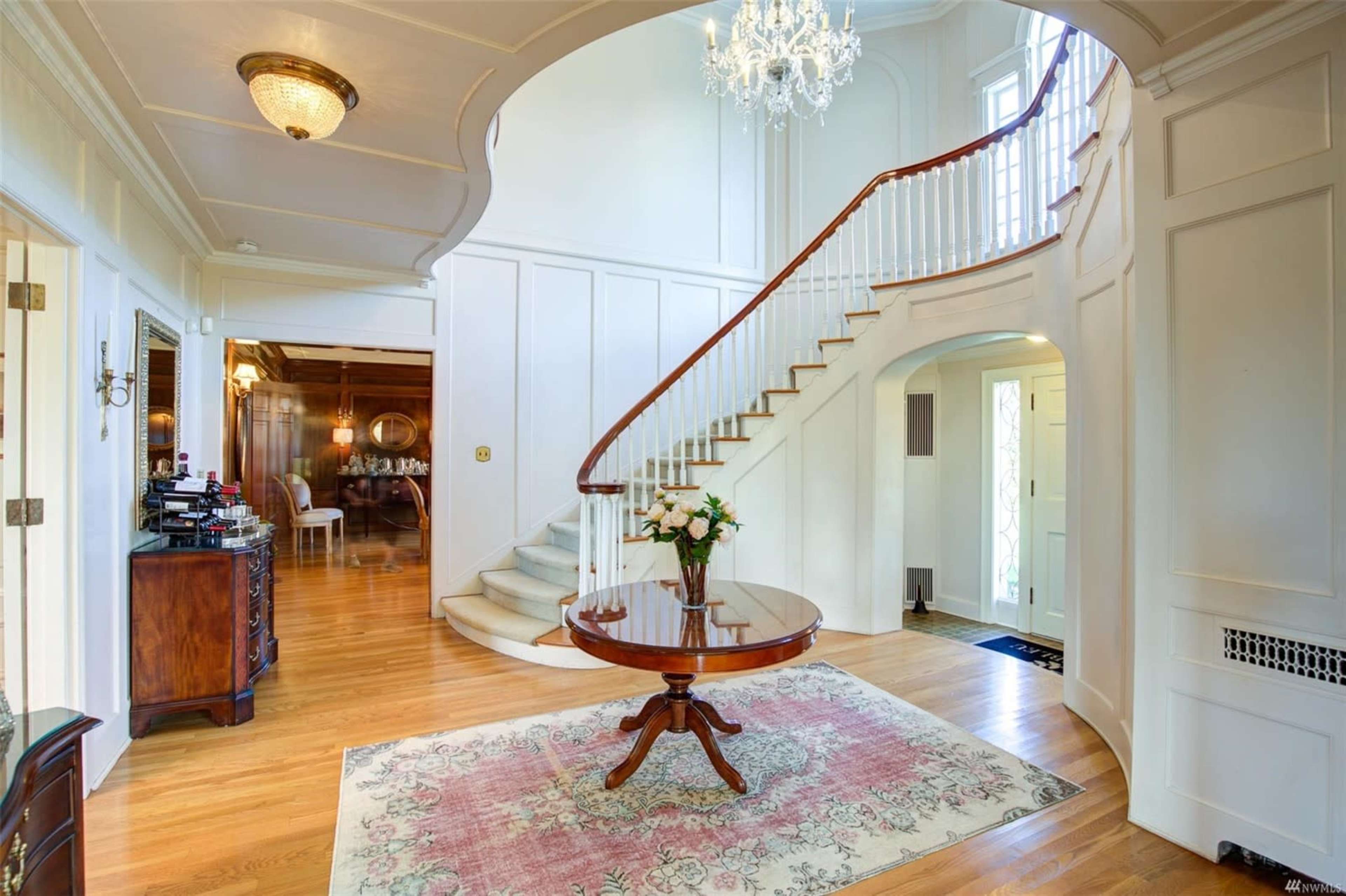 The image shows a brightly lit entryway with a curved staircase, an ornate chandelier, and a round table with flowers on a patterned rug.
