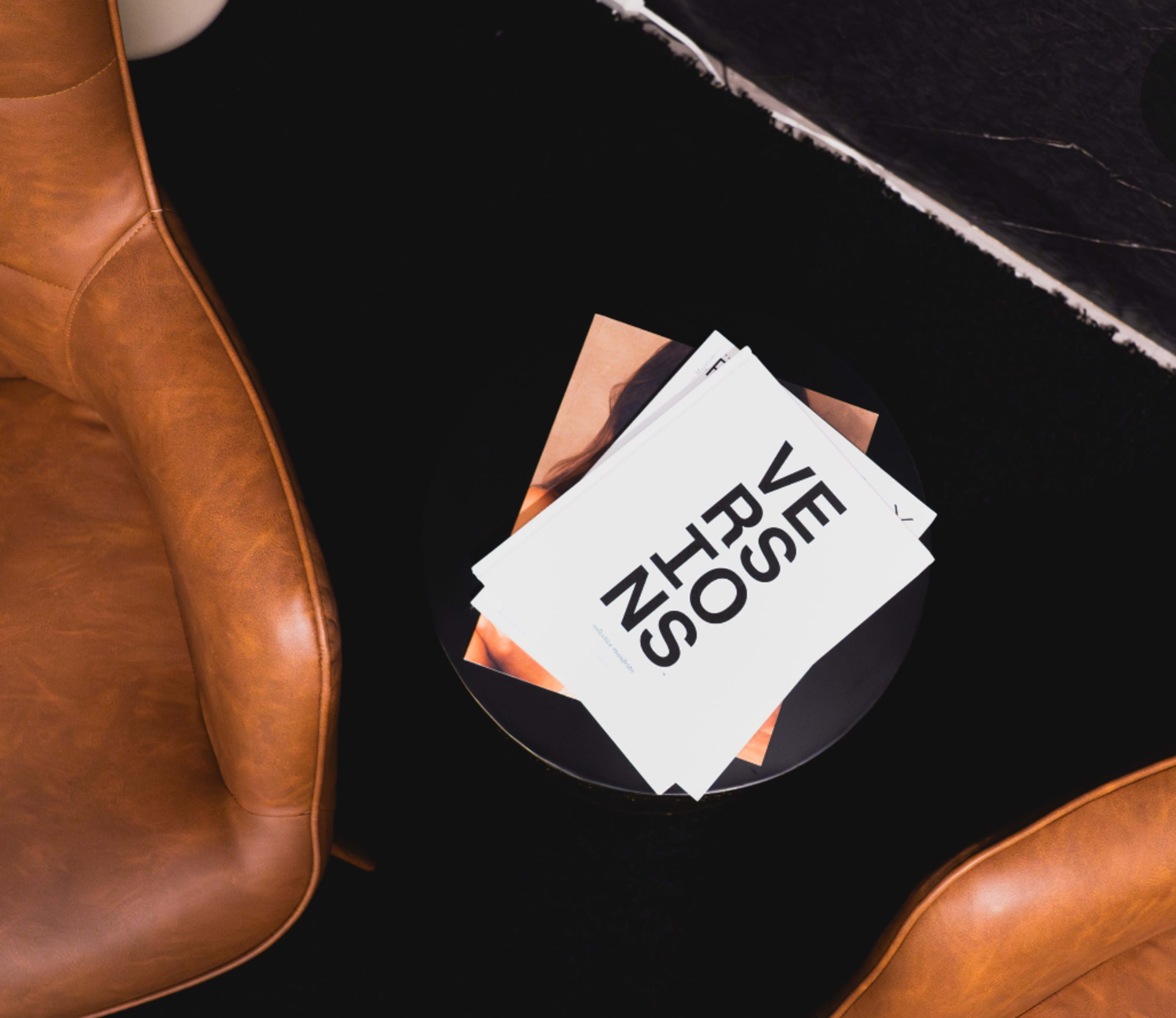 Two brown leather chairs are positioned beside a small black table holding several magazines with bold text on the cover.