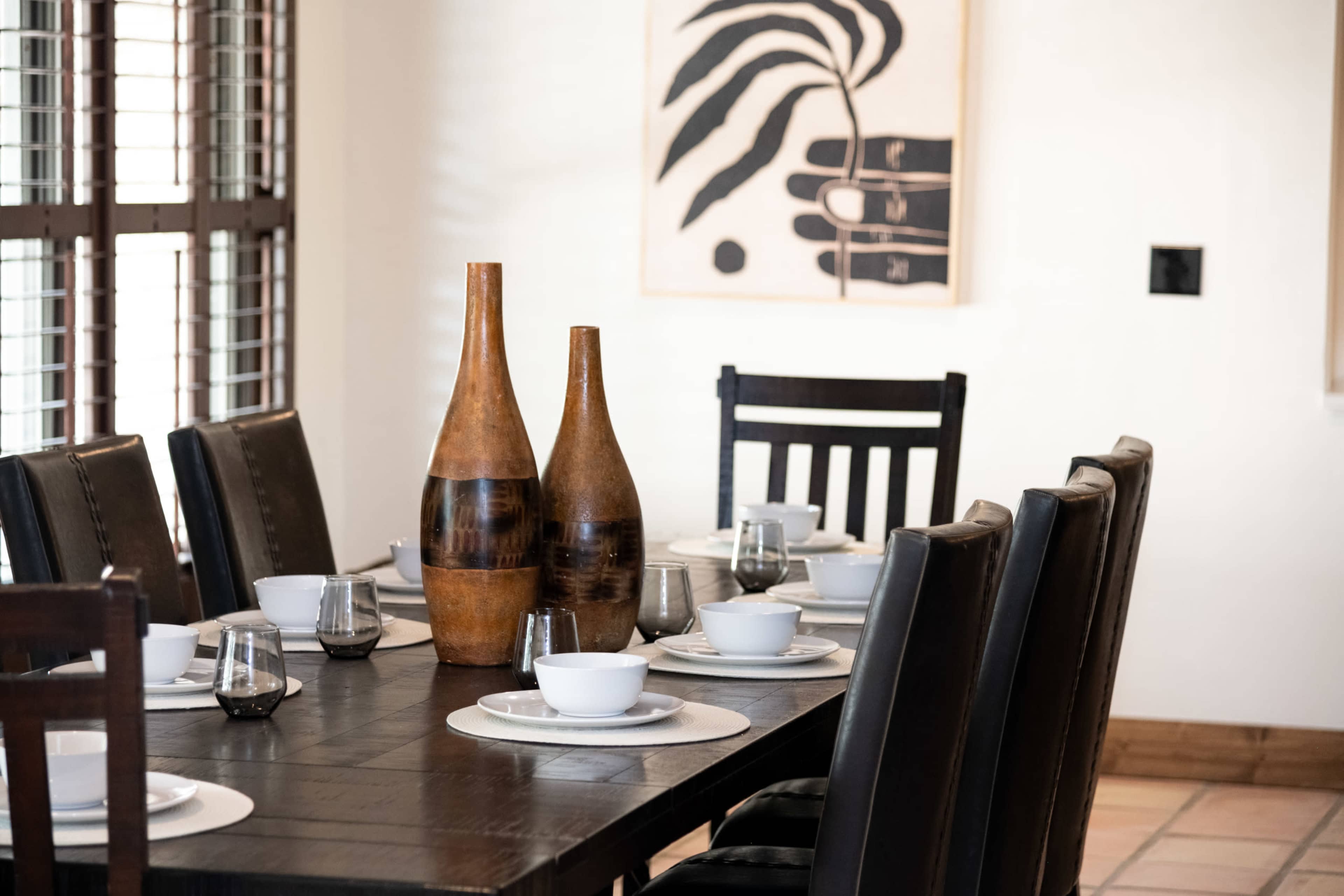 A wooden dining table is set with white bowls and glassware, flanked by black chairs and adorned with two decorative vases, against a wall featuring a piece of artwork.