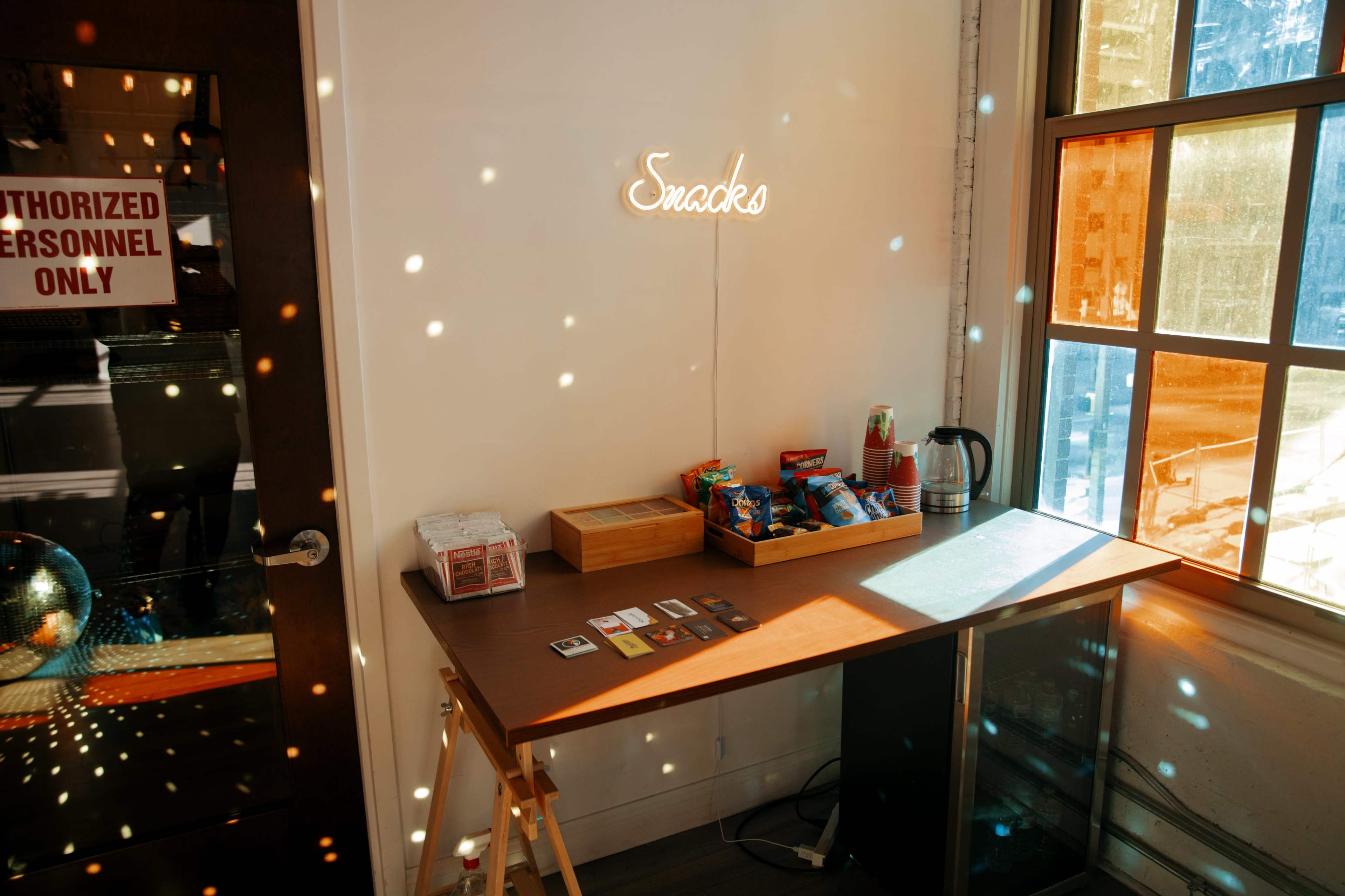 A table with snacks and a kettle is positioned against a wall with a neon sign that says "Snacks" and a door labeled "Authorized Personnel Only."