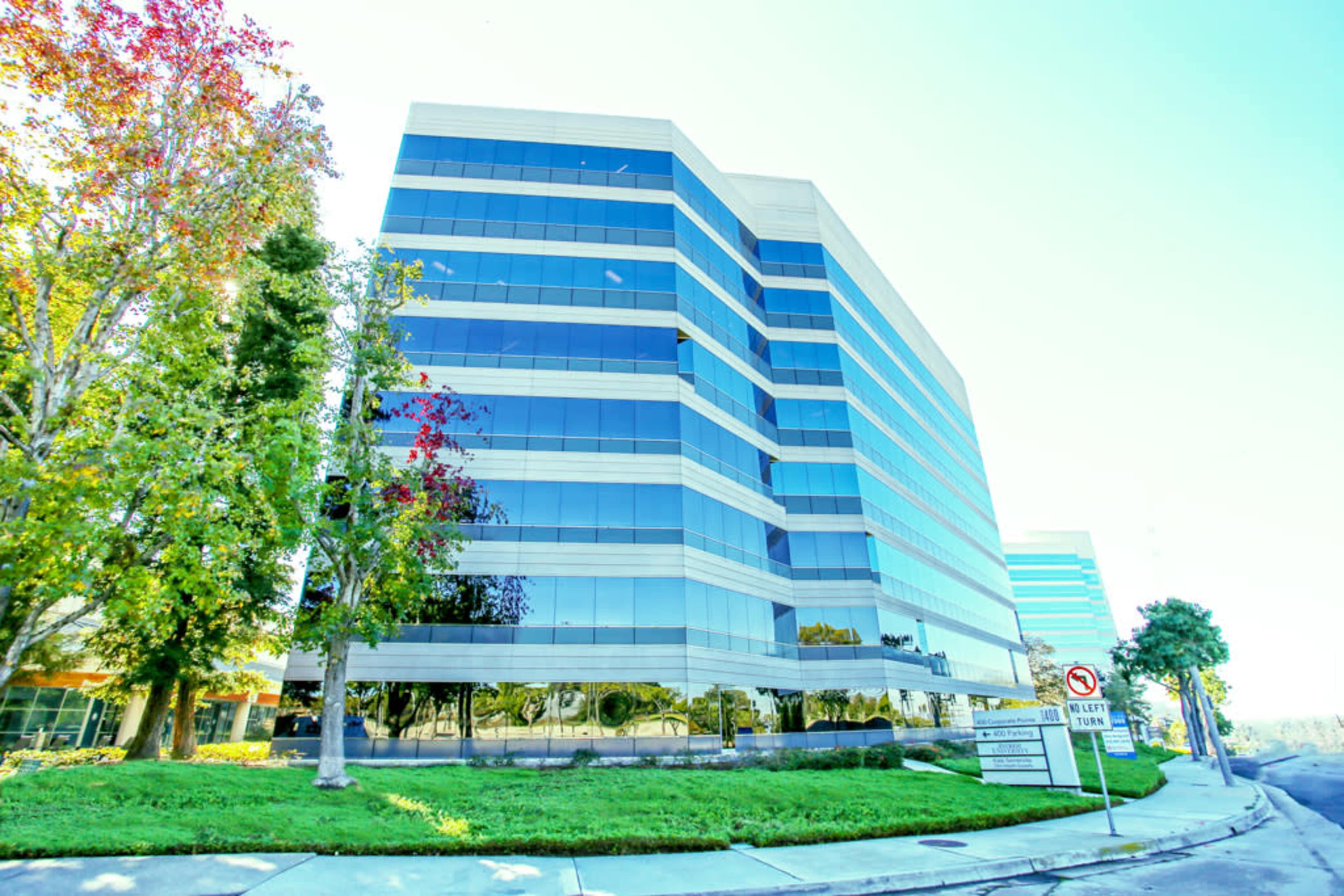 A modern multi-story office building with large glass windows is surrounded by well-maintained landscaping and trees.