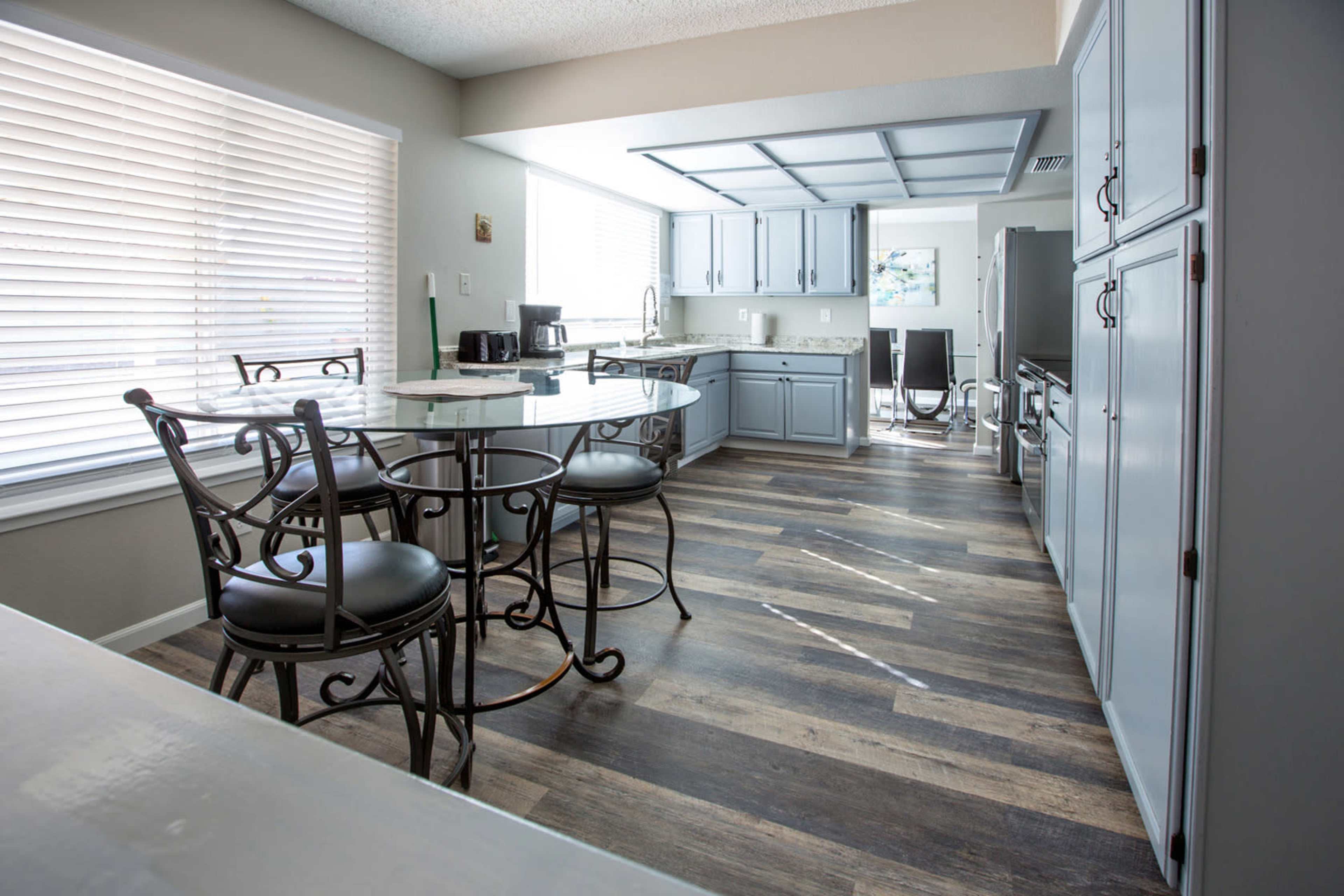A modern kitchen features gray cabinetry, a central island with bar stools, and a dining area illuminated by natural light through the windows.