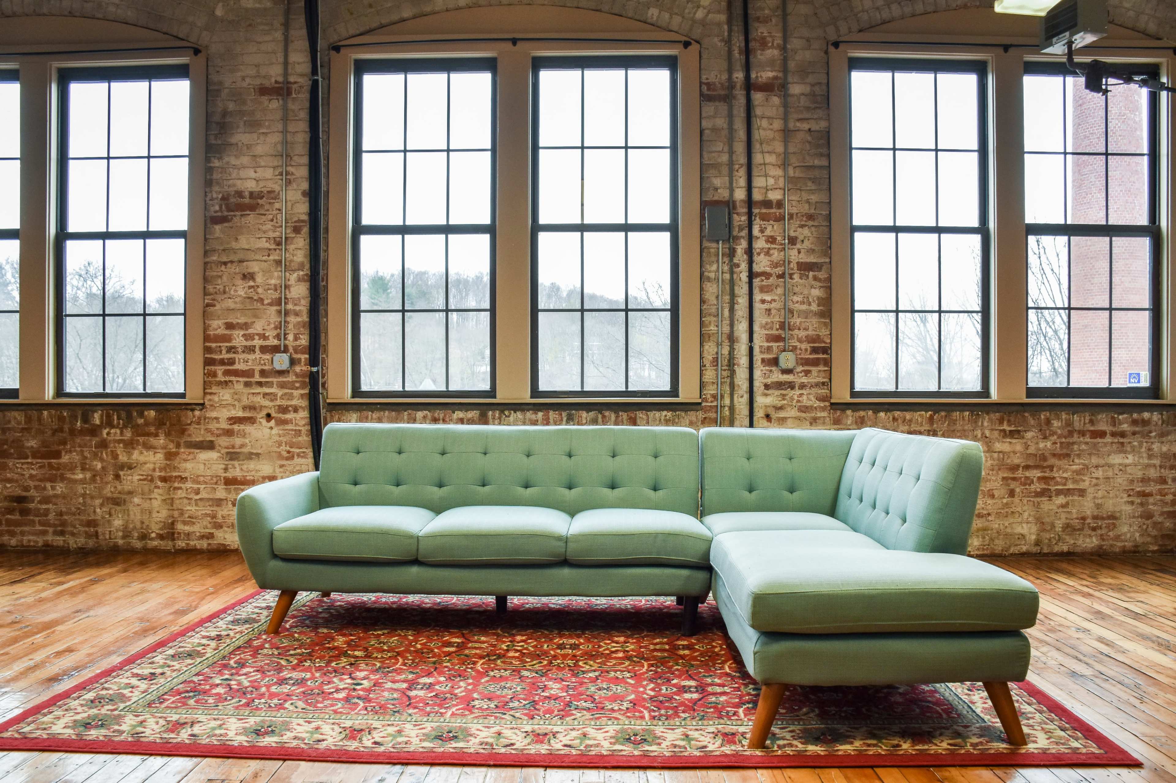 A light green sectional sofa is positioned on a patterned rug in a room with large brick walls and multiple windows.