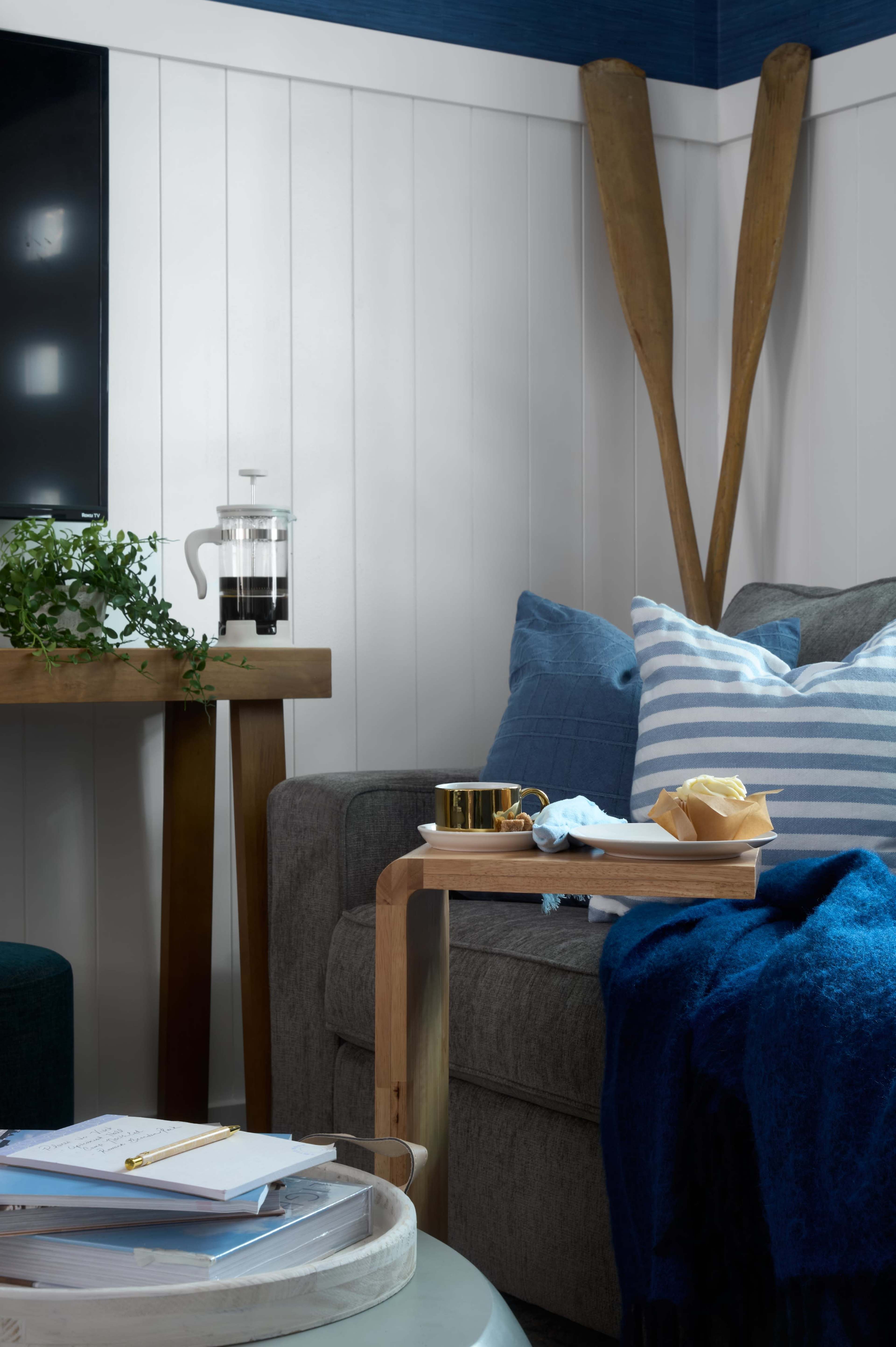 A gray sofa with blue and white striped cushions is positioned beside a wooden table holding a French press and a gold cup, while a blue blanket drapes over the arm of the sofa.