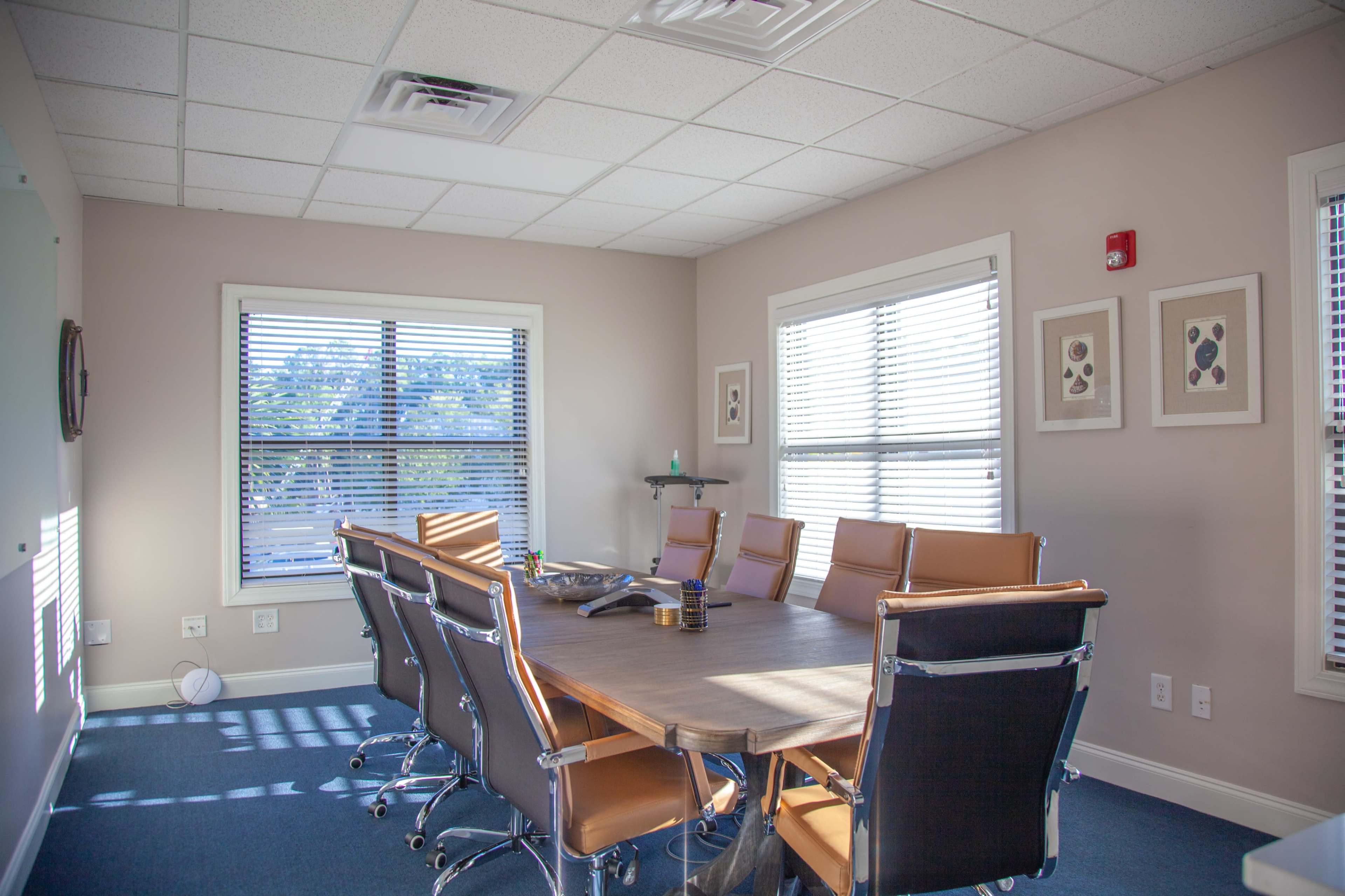A conference room features a large wooden table surrounded by eight leather chairs and is illuminated by natural light through two windows.