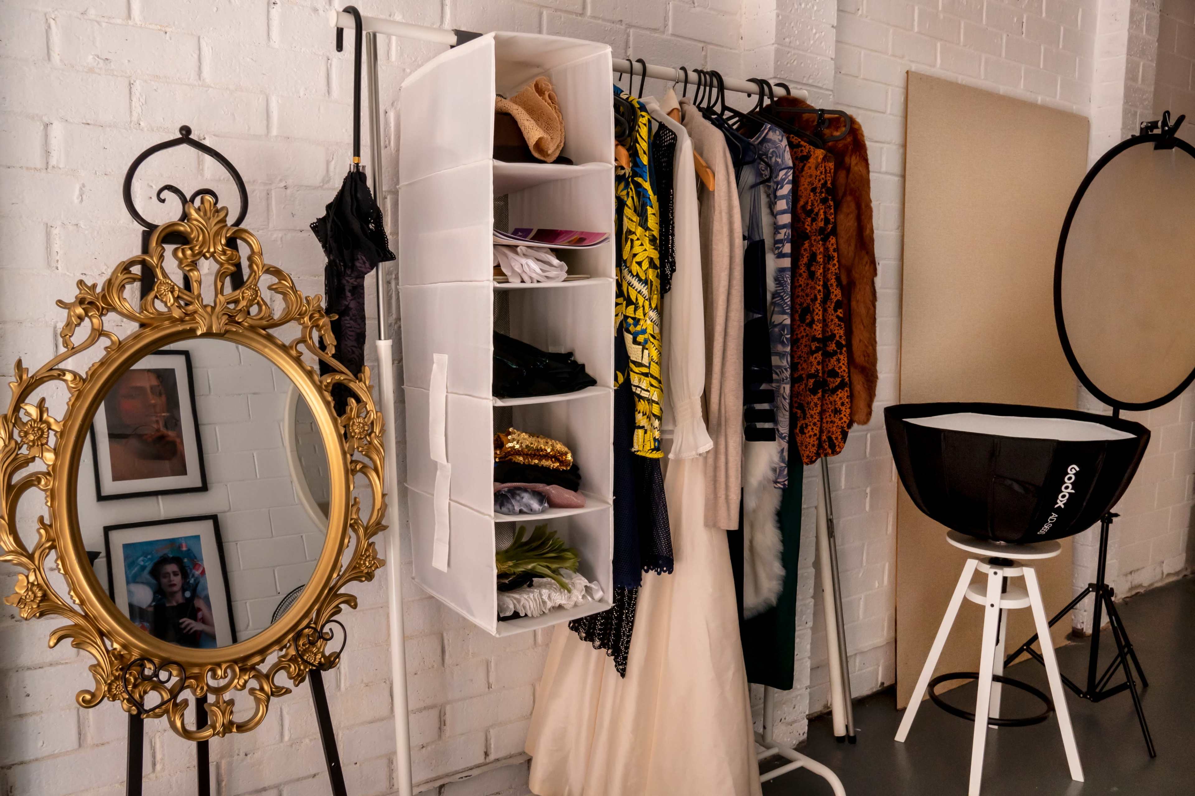 A clothing rack with various garments, a hanging organizer with folded items, a decorative mirror, and a photography light setup against a white brick wall.