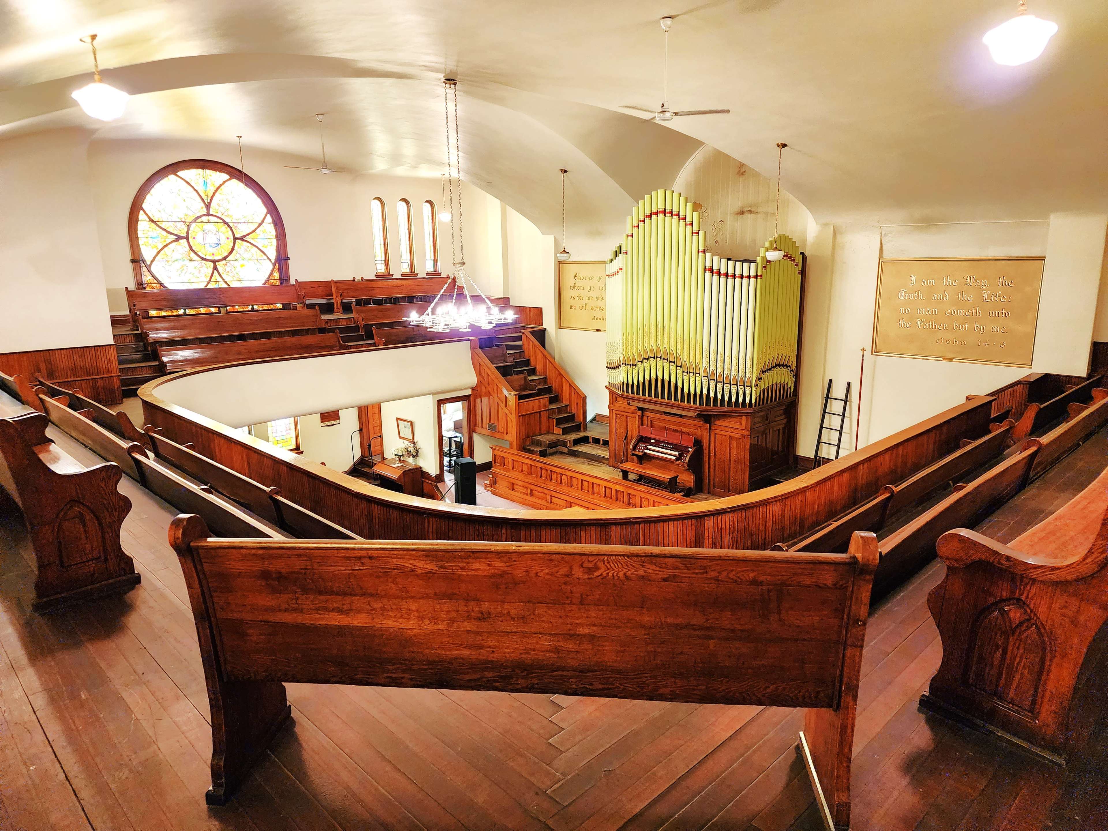 The interior of a church featuring wooden pews, an organ prominently displayed at the front, and a large stained glass window.