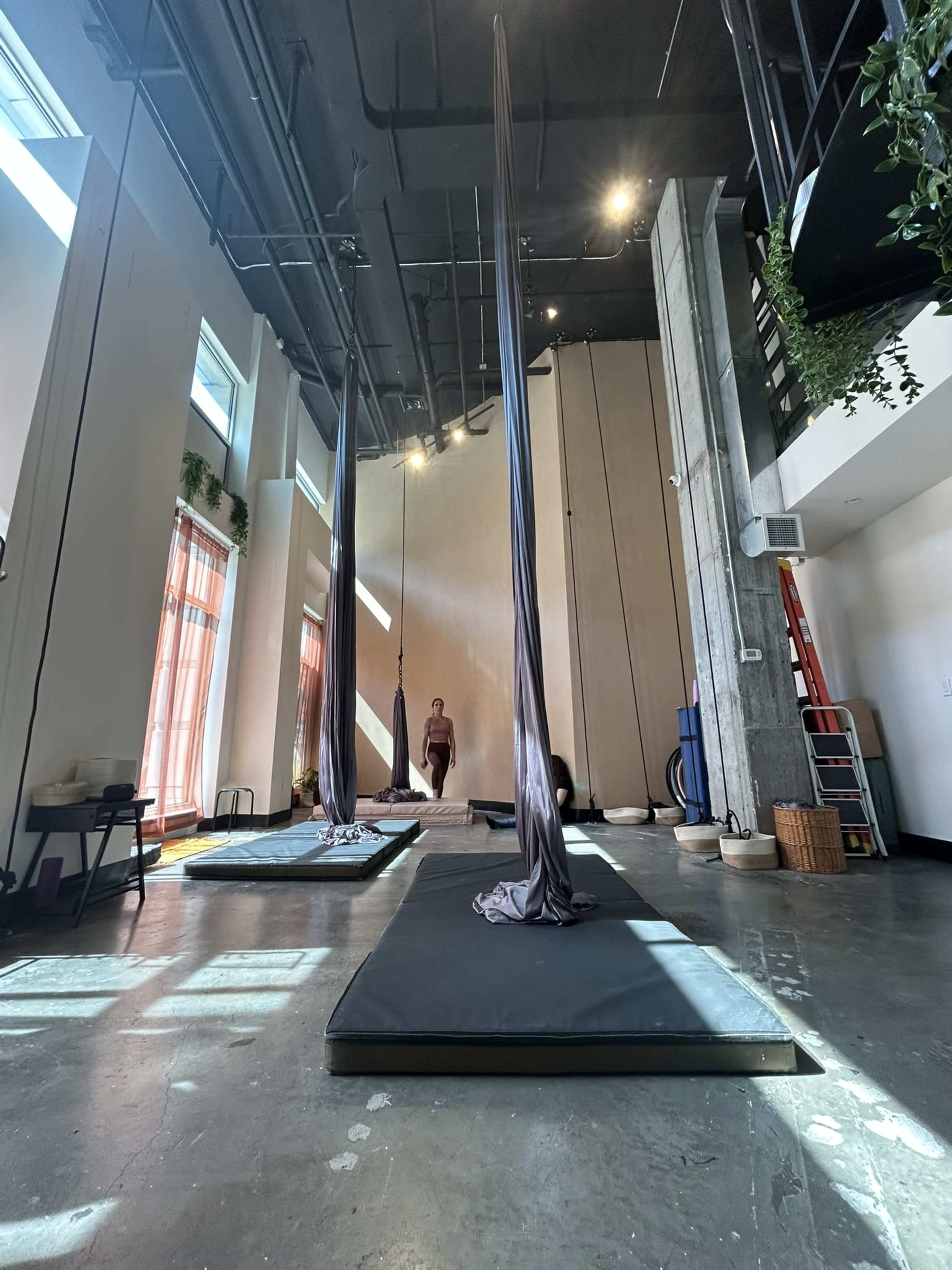 A spacious studio with high ceilings features two aerial silk apparatus suspended from the ceiling and a person standing on a black mat in the center.
