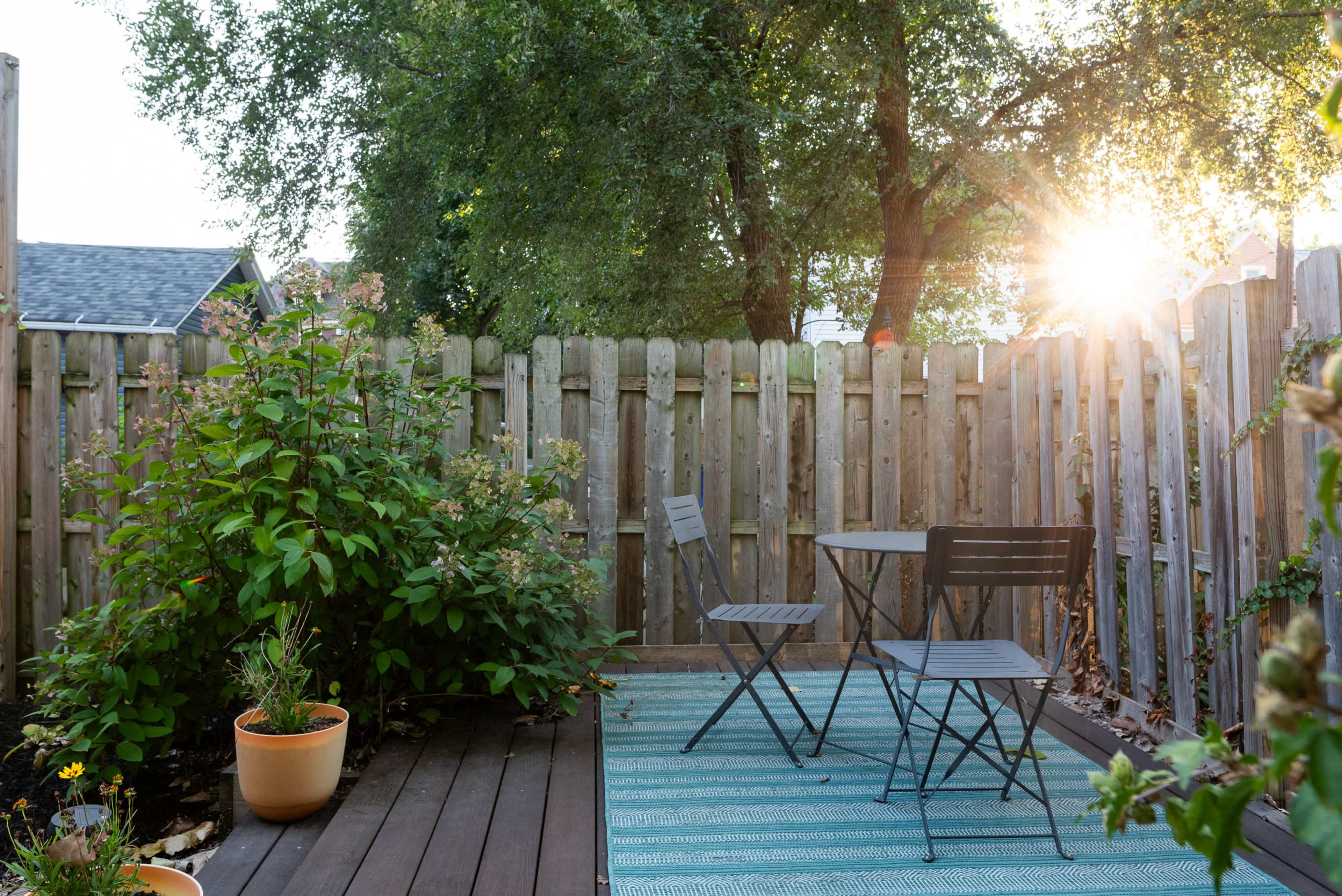 A small patio area features two folding chairs and a table surrounded by garden plants and a wooden fence, with the sun setting in the background.