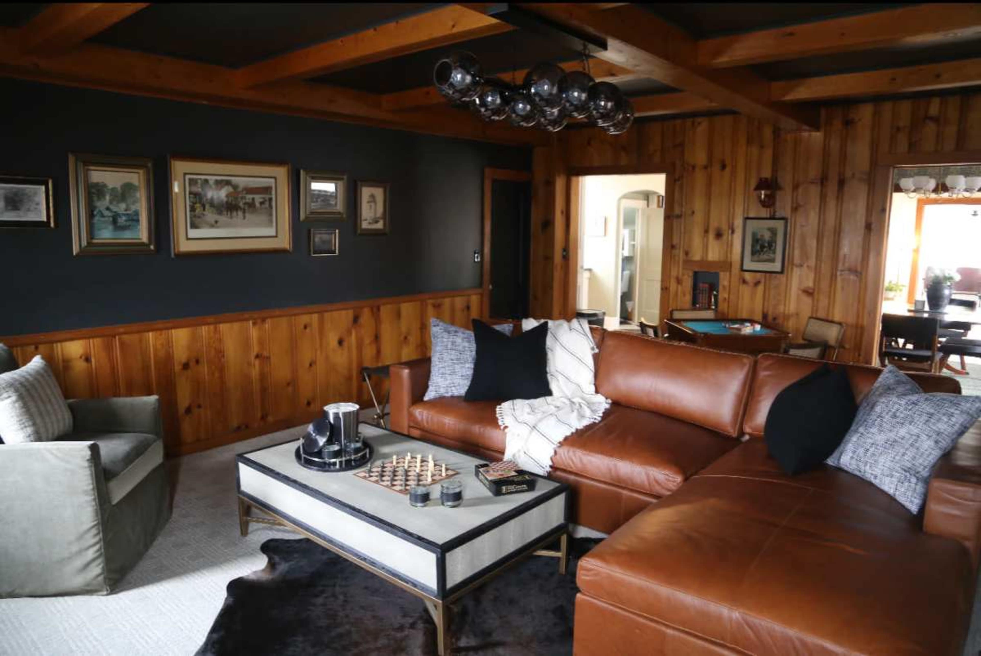 The image shows a cozy living room with wooden paneling, a brown leather sectional sofa, a glass coffee table, and decorative items including a chess set and wall art.