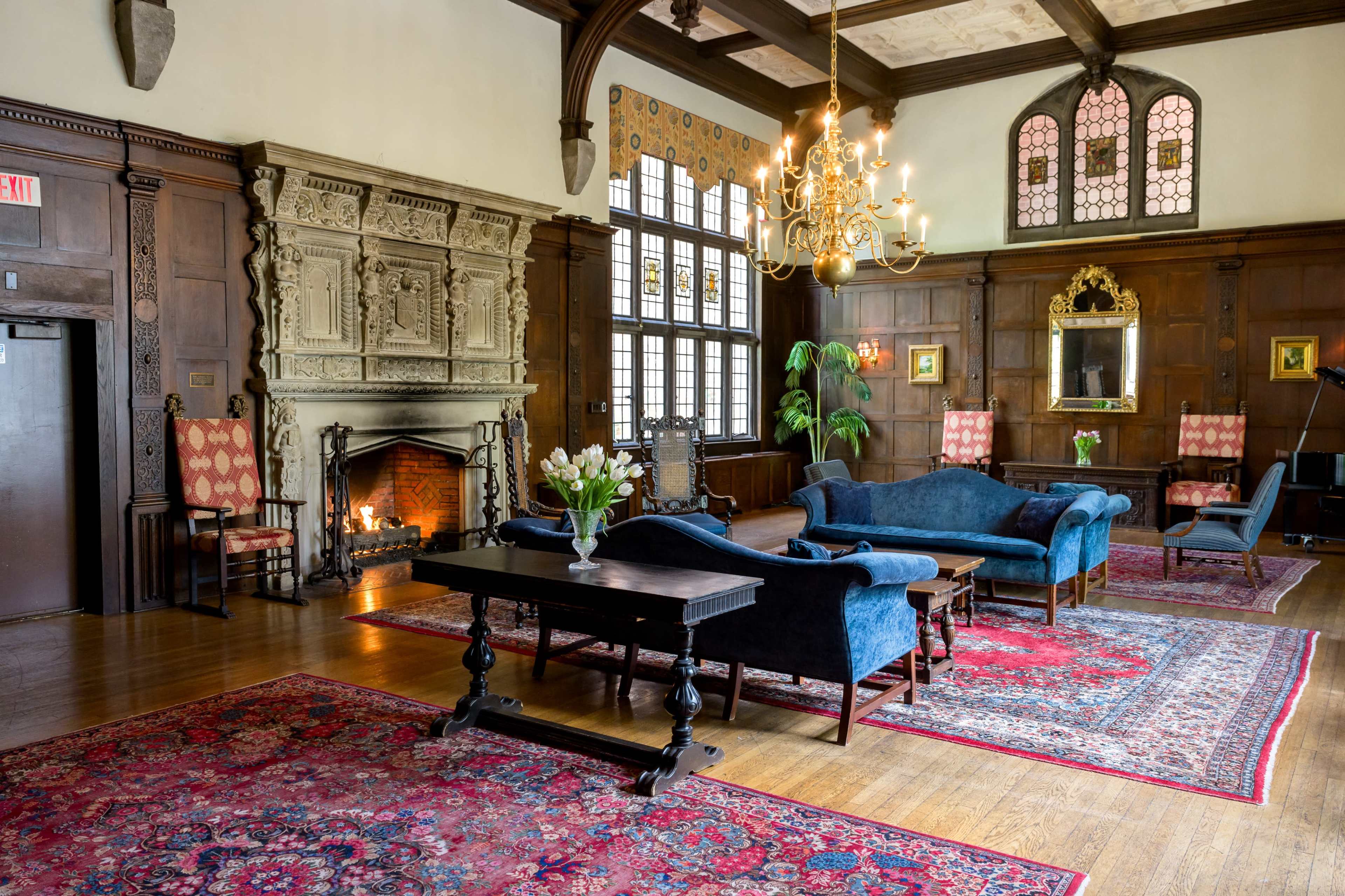 An elegant room with wooden walls, a large fireplace, a chandelier, and a mix of blue upholstered furniture and decorative rugs.