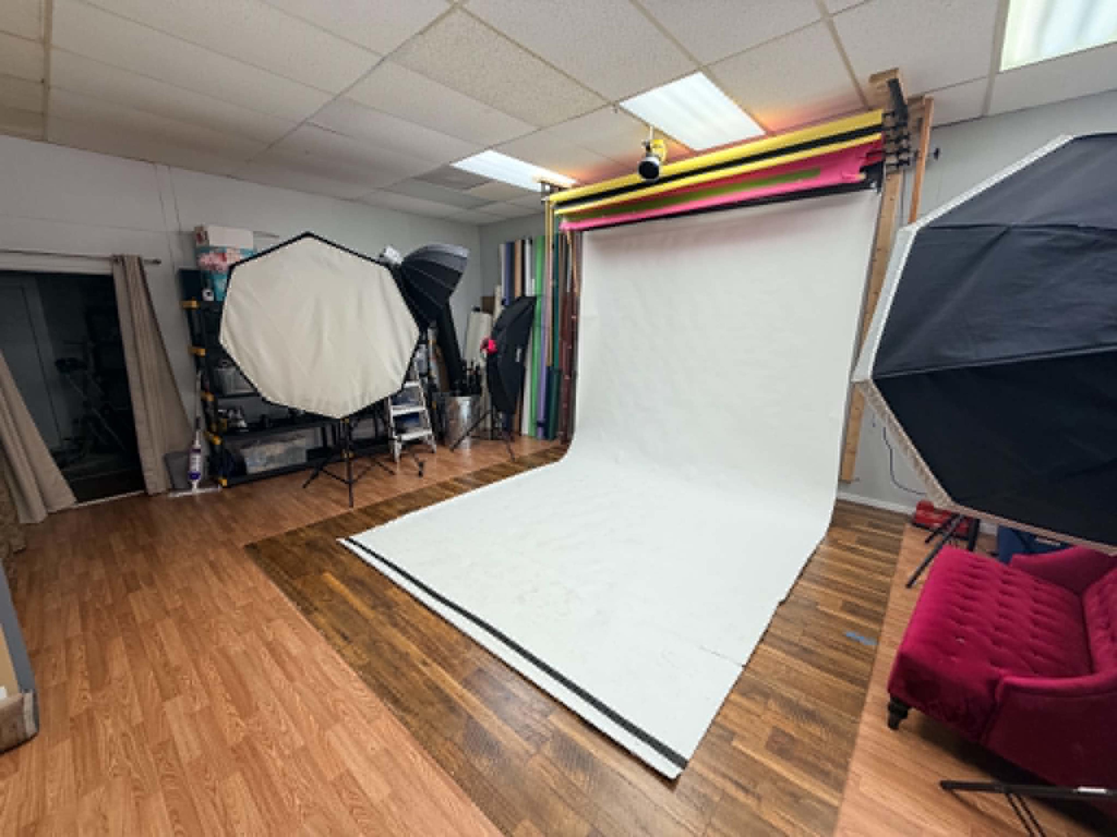 The image shows a photography studio with a white backdrop, softbox lighting setups, and wooden flooring.