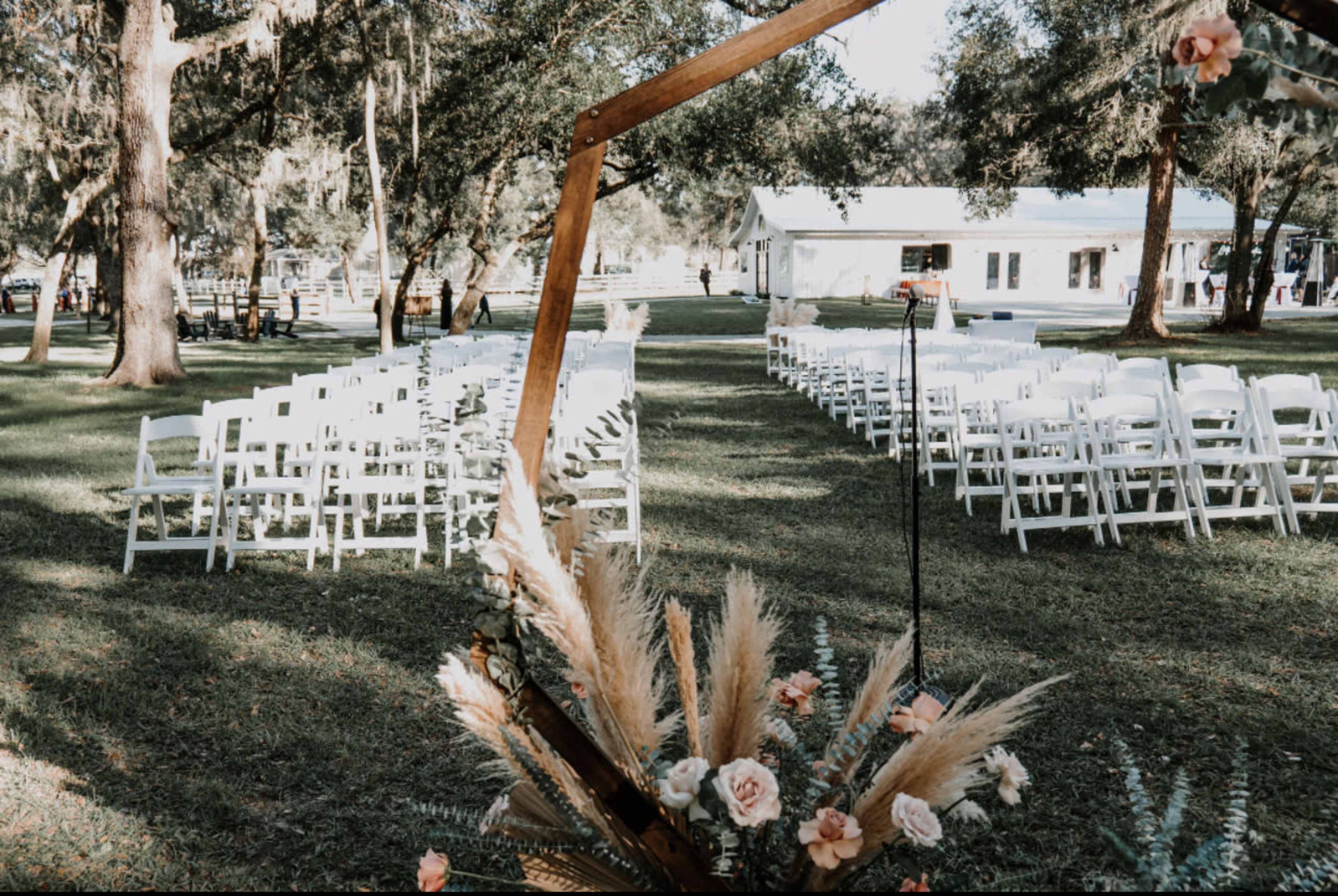 An outdoor wedding ceremony setup with white chairs arranged in rows under trees, leading to an archway decorated with floral elements.