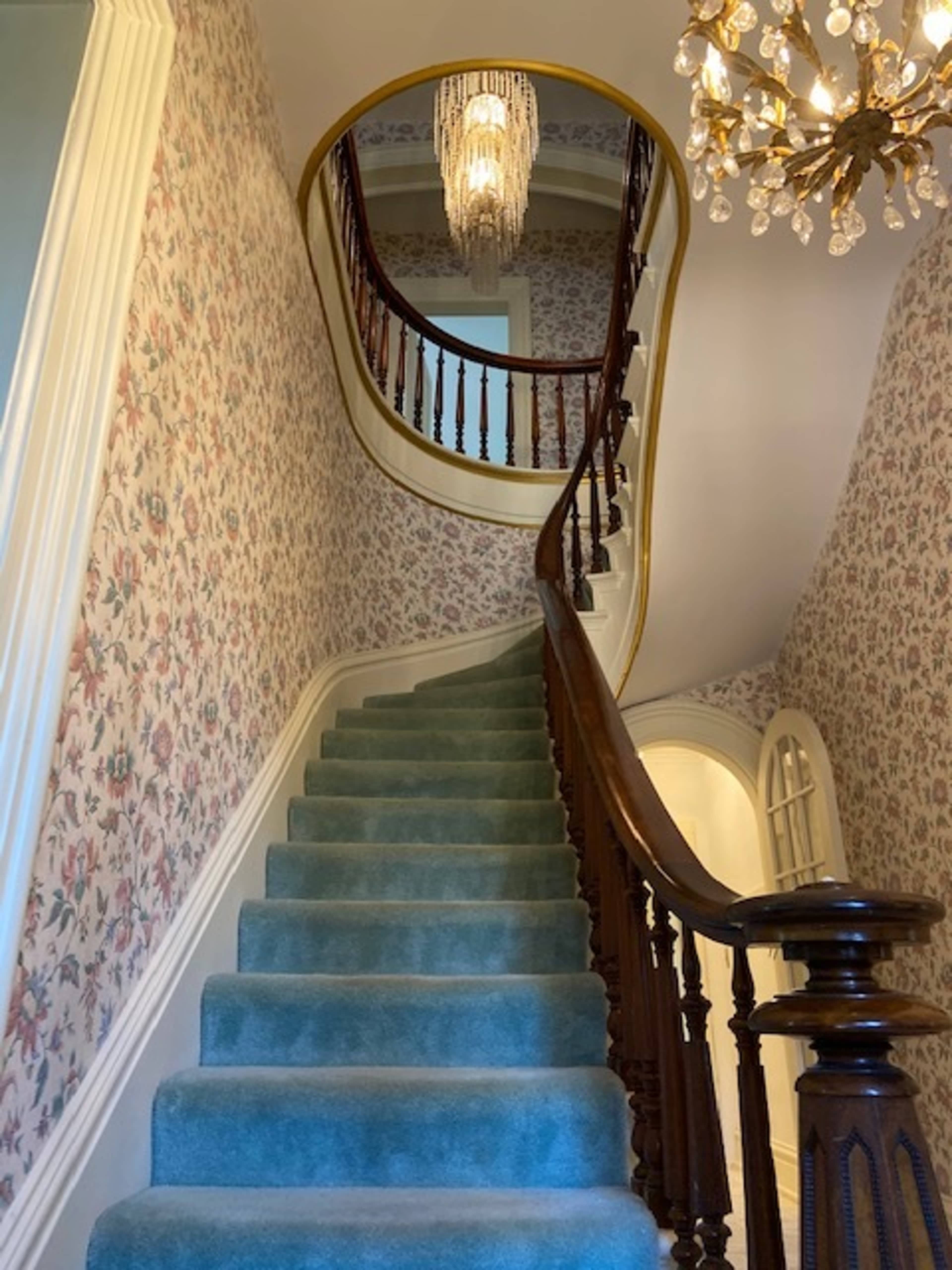 A winding staircase with blue carpet leads up to a chandelier, set against floral wallpaper.