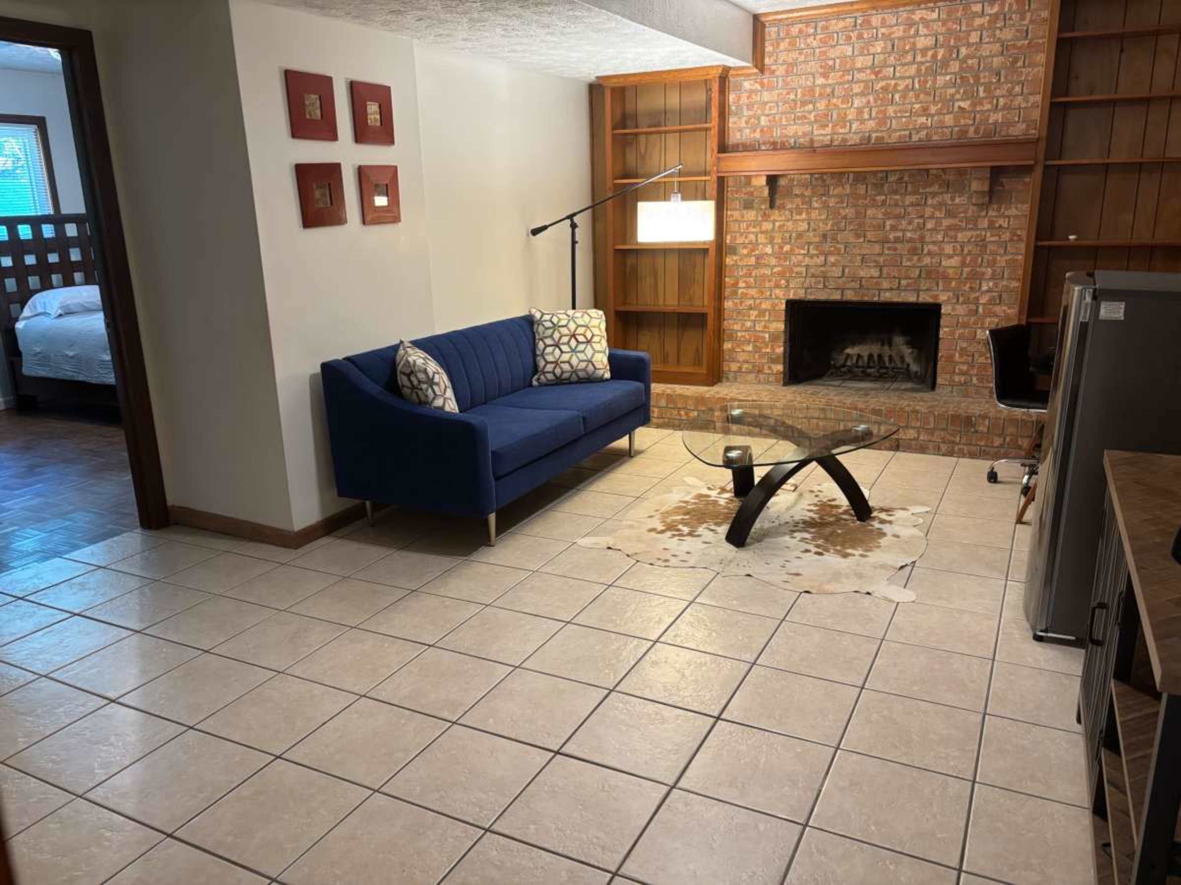 The image shows a living room with a blue couch, a glass coffee table, and a brick fireplace, with tiled flooring and wood furnishings in the background.