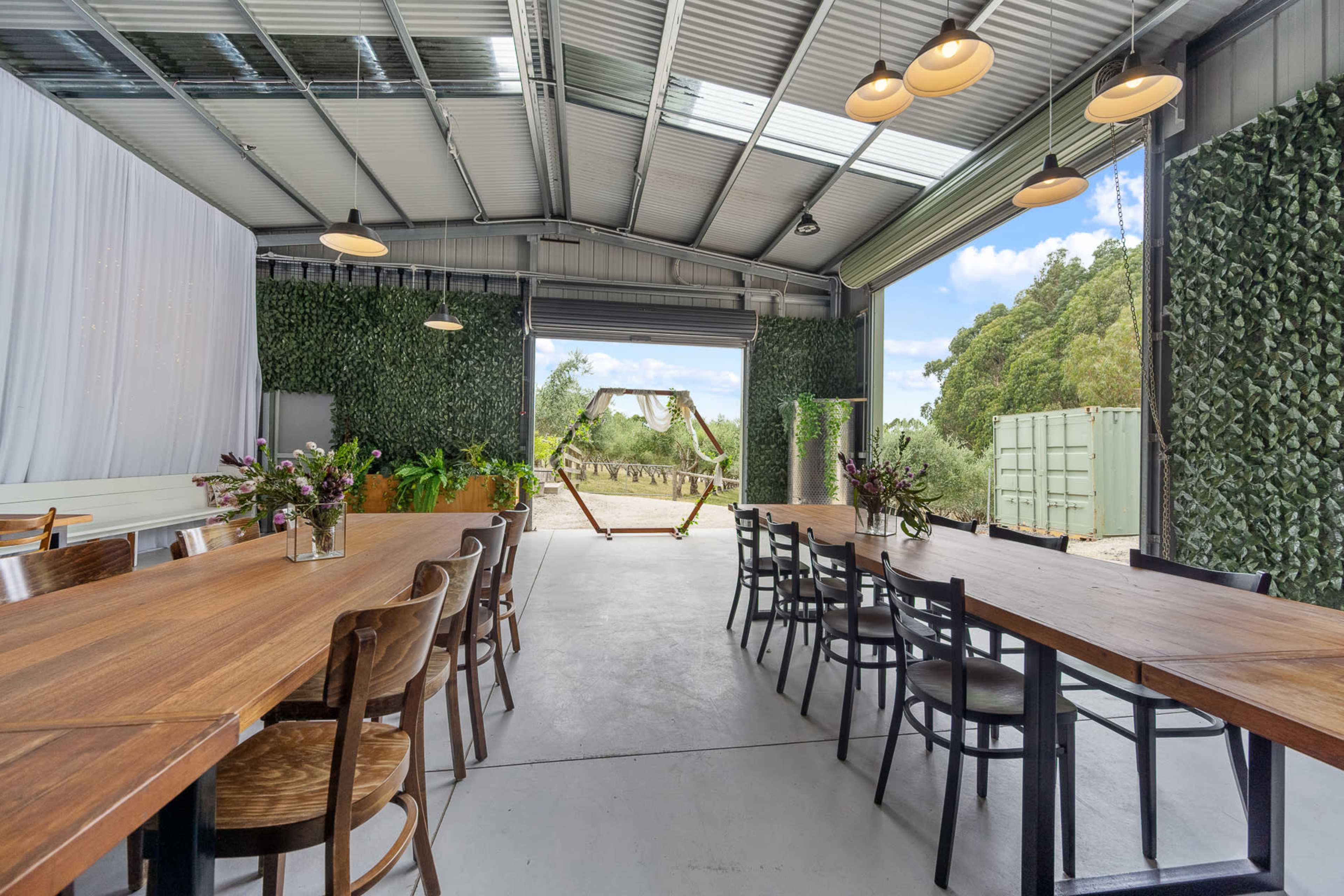 The image shows a spacious indoor area with wooden tables and chairs, featuring greenery on the walls and an open view of a swing set outside.
