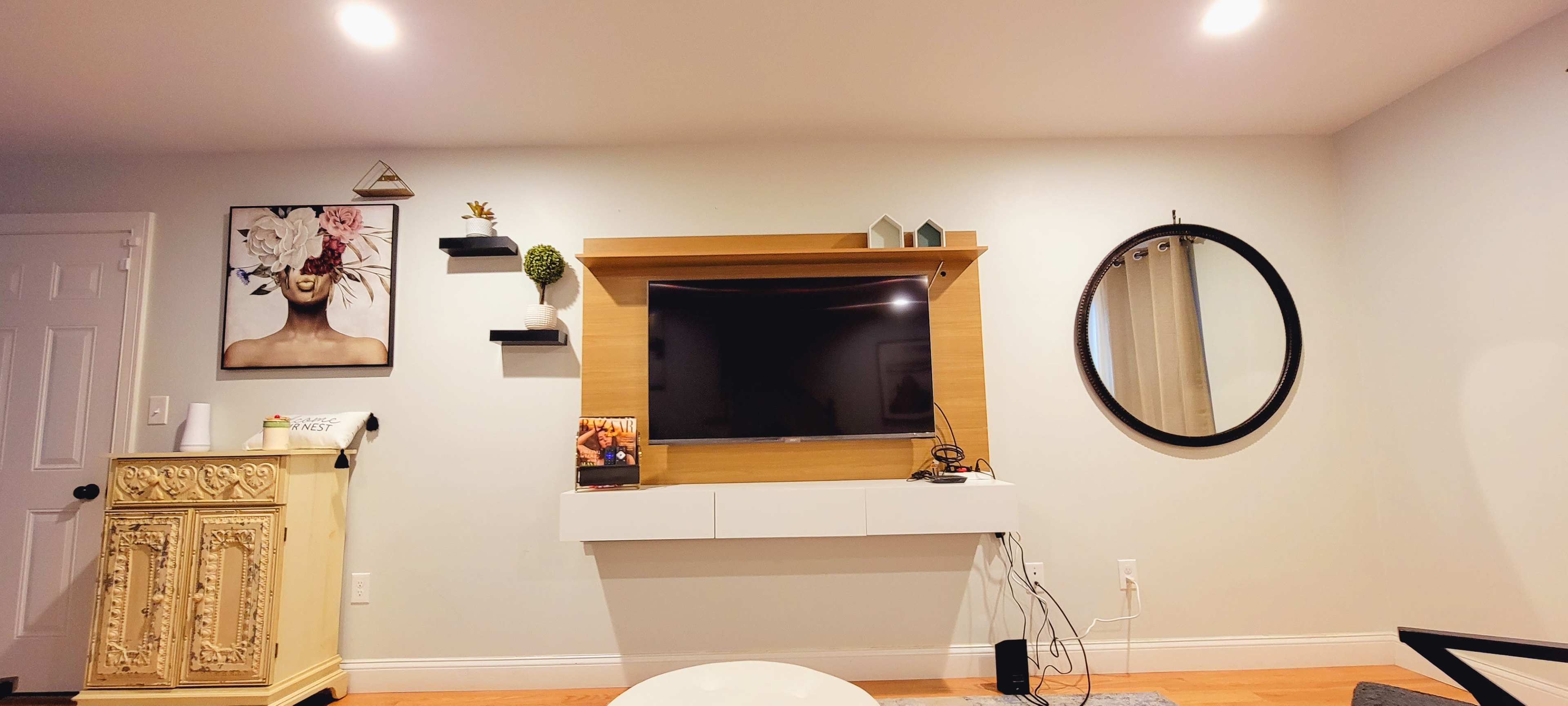 A living room with a mounted flat-screen TV above a wooden media console, surrounded by decorative shelves and a round wall mirror.