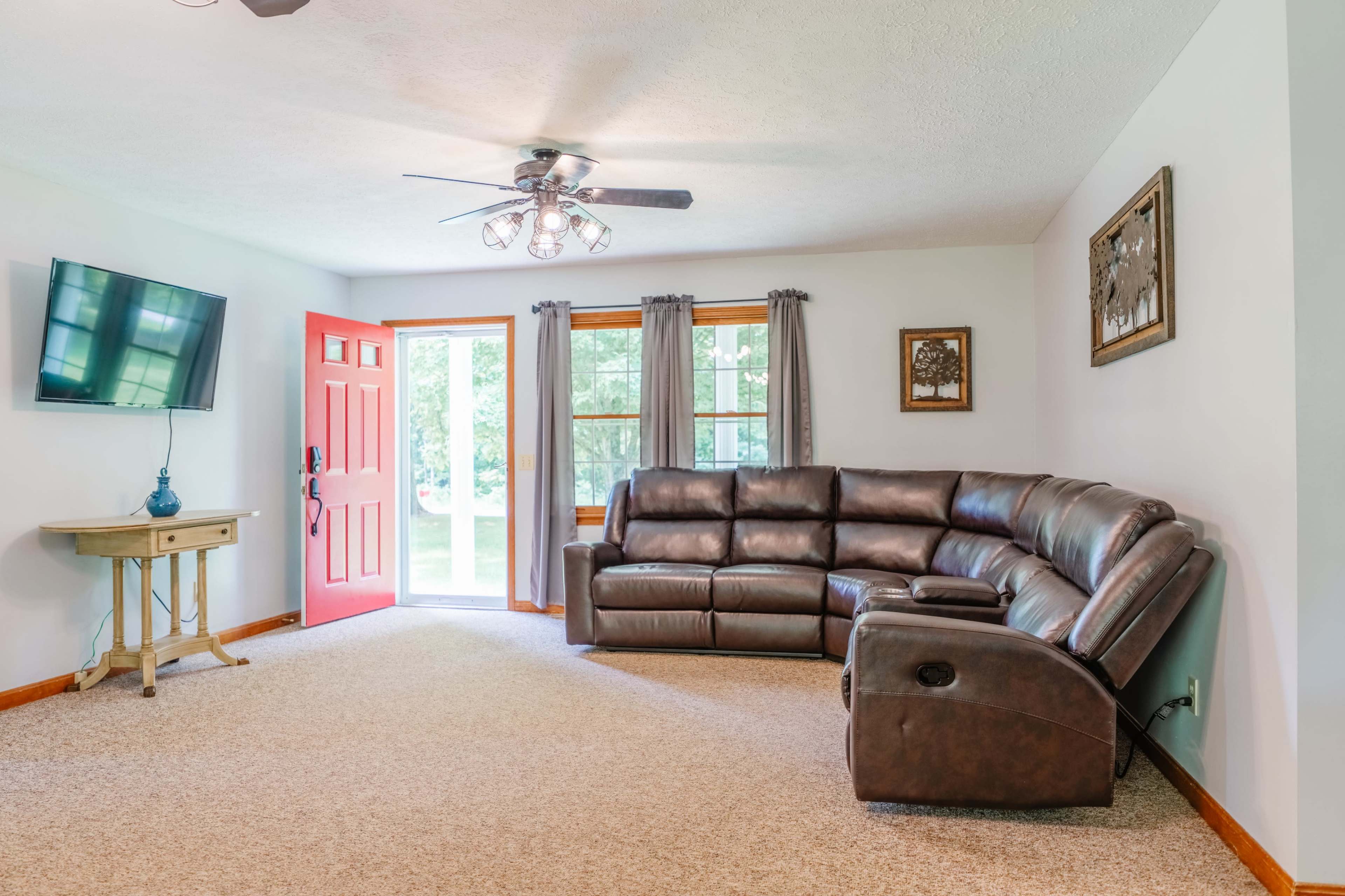 The image shows a living room featuring a brown sectional sofa, a television mounted on the wall, and a bright red front door leading to an outdoor view.