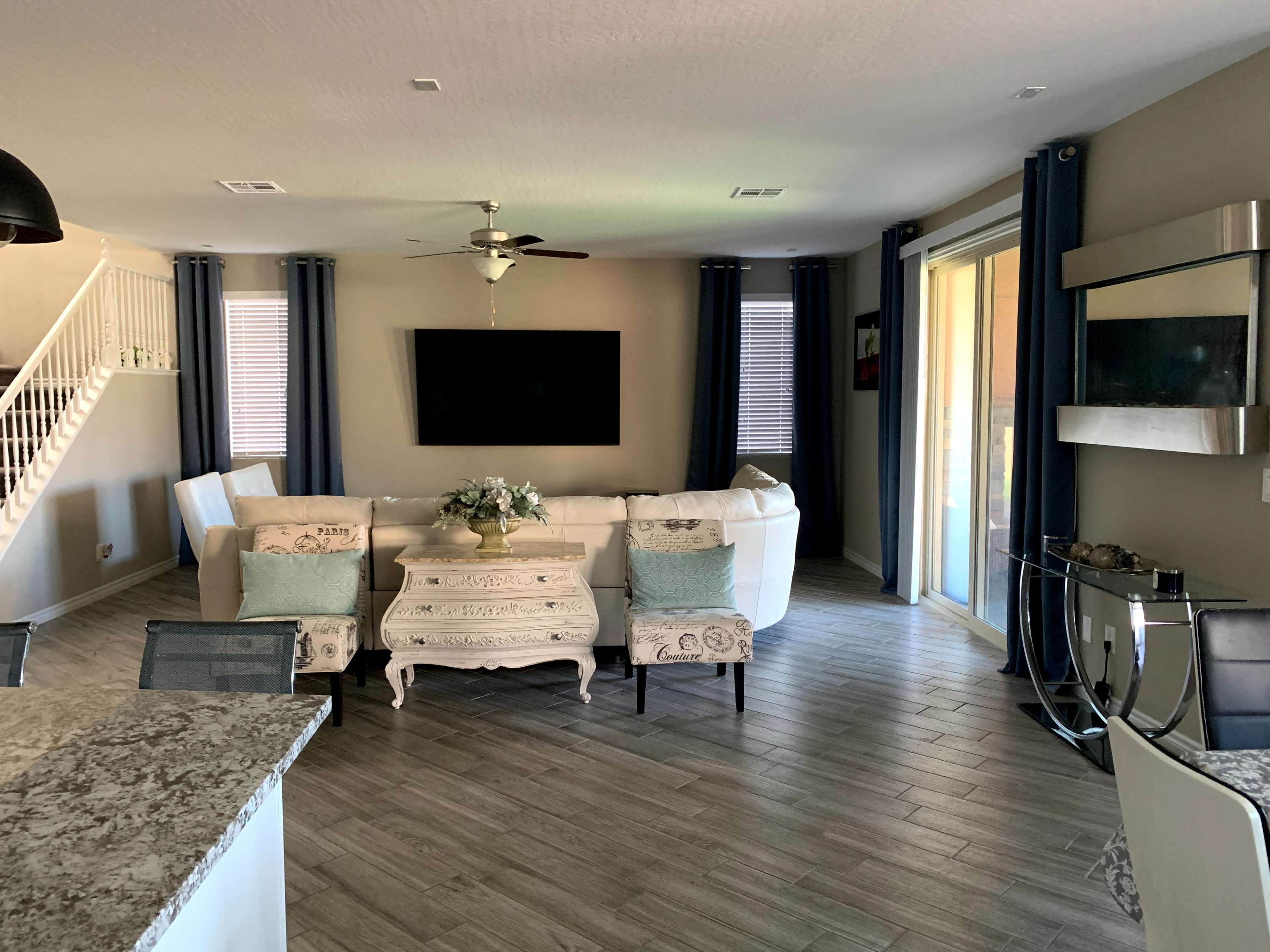 A living room features a white sectional sofa, a mounted TV, a coffee table, and large windows with blue curtains.