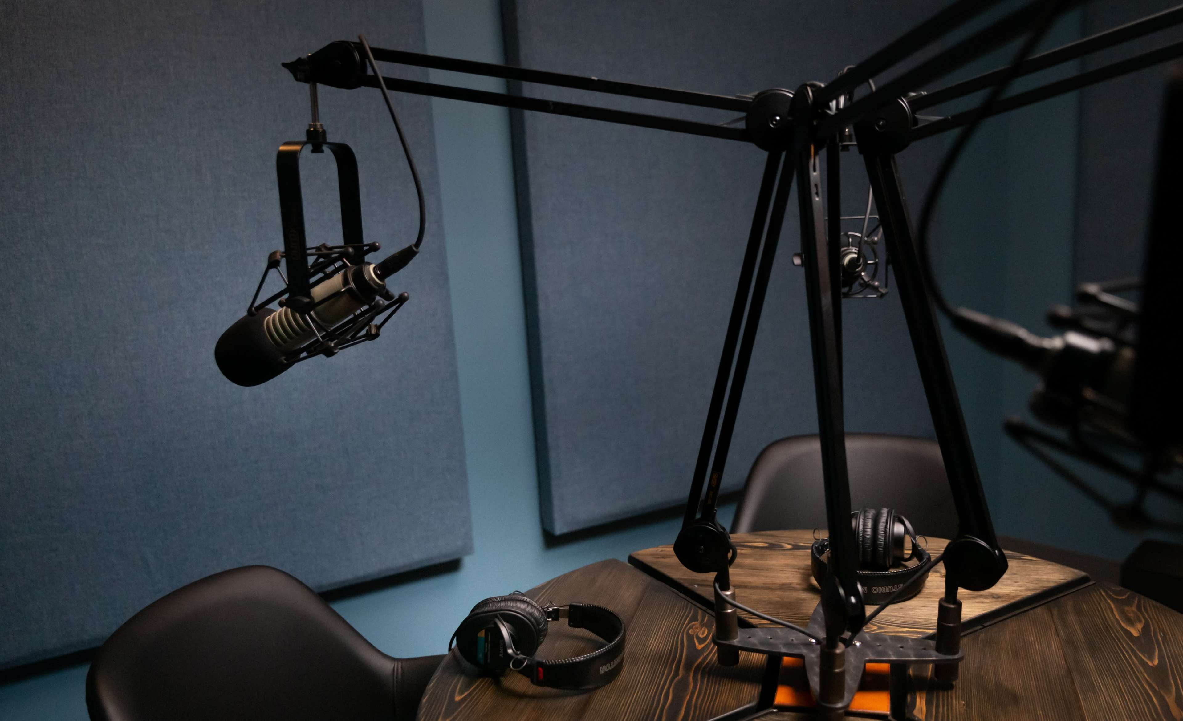 The image shows a podcast recording setup with two microphones on adjustable arms and headphones placed on a wooden table, surrounded by soundproofing panels.