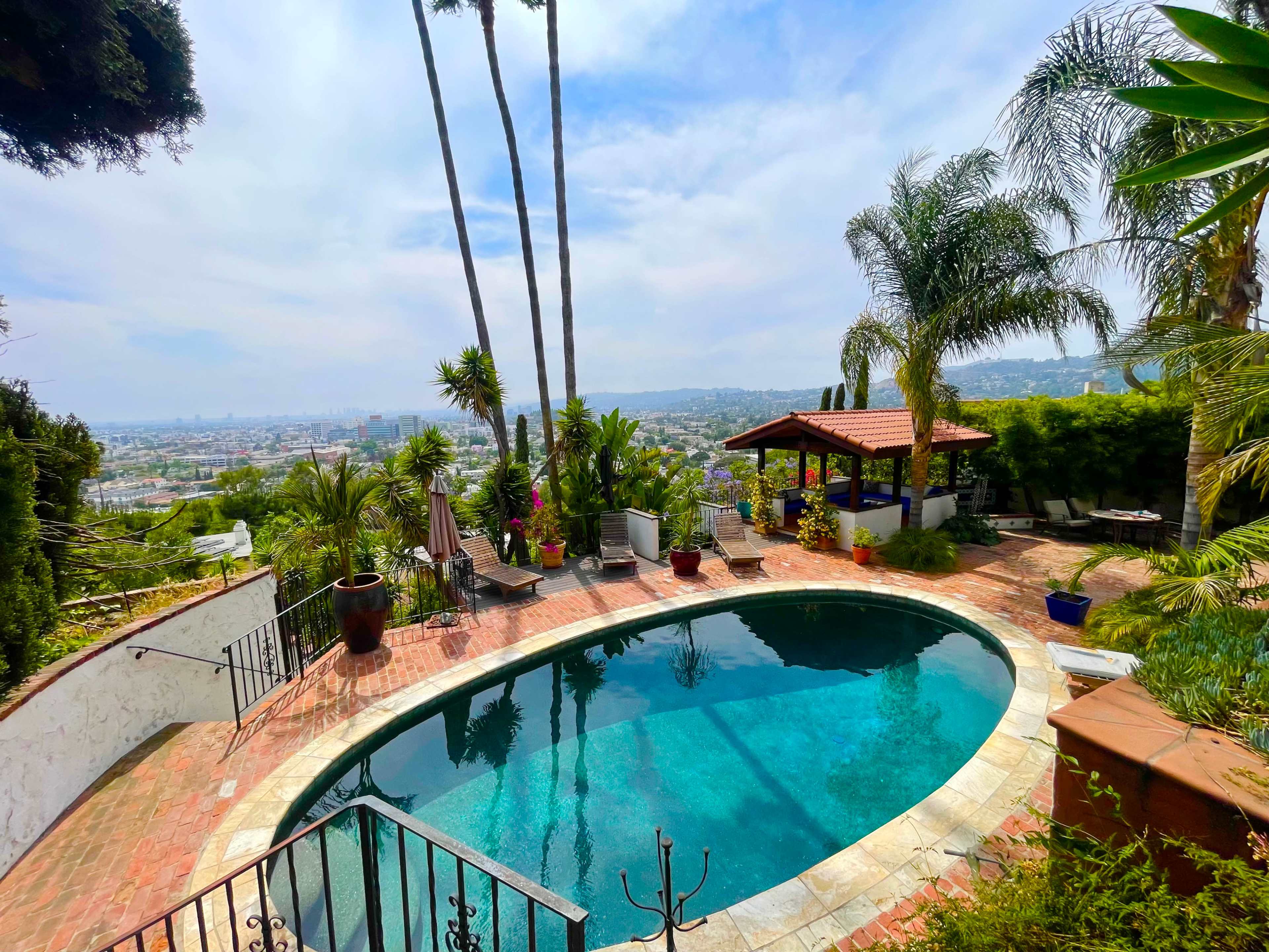 The image shows a curved swimming pool surrounded by lush greenery, with a view of a city in the distance and a shaded seating area nearby.
