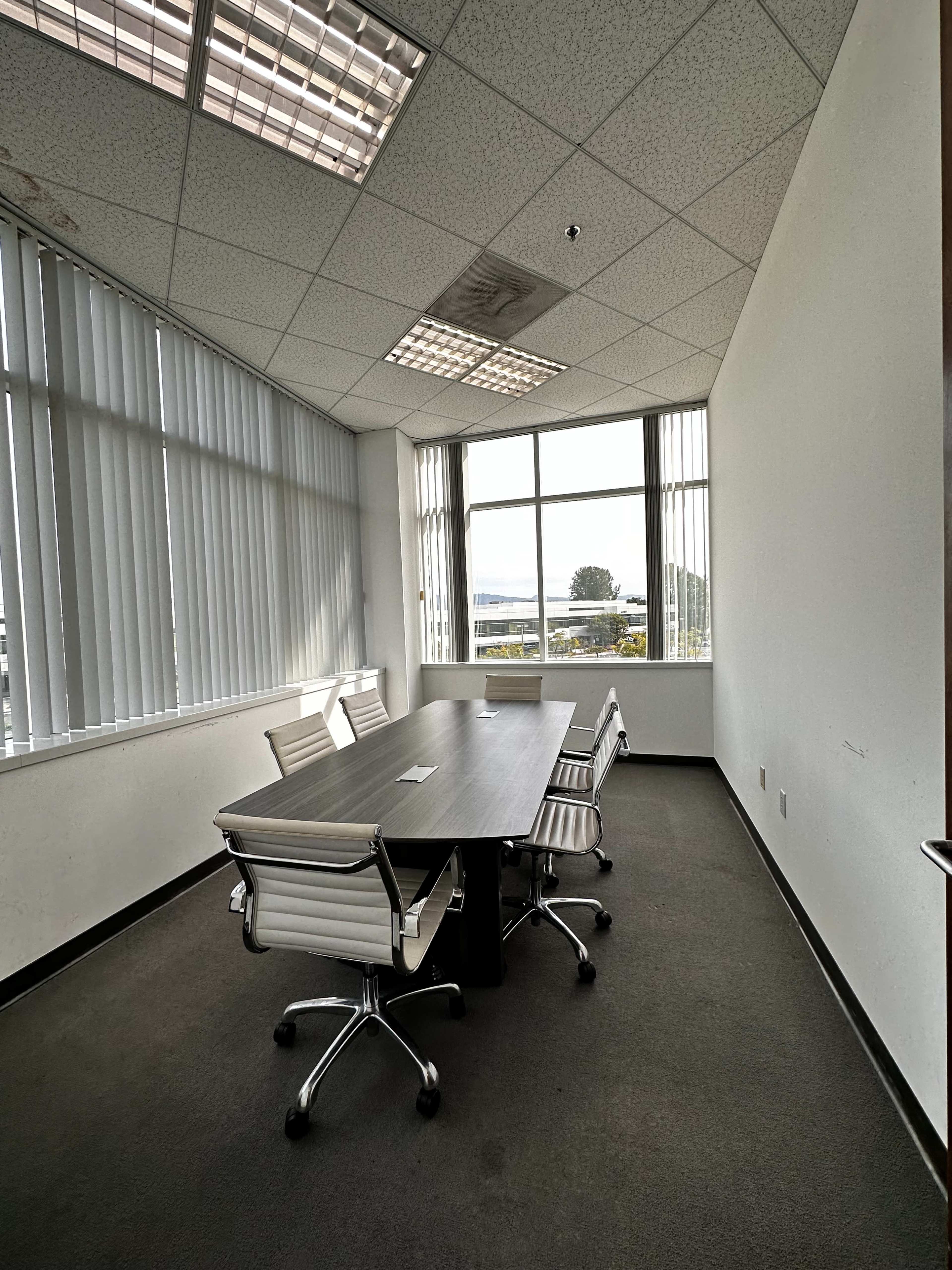 A small conference room features a rectangular table surrounded by four white office chairs, with large windows allowing natural light to enter.