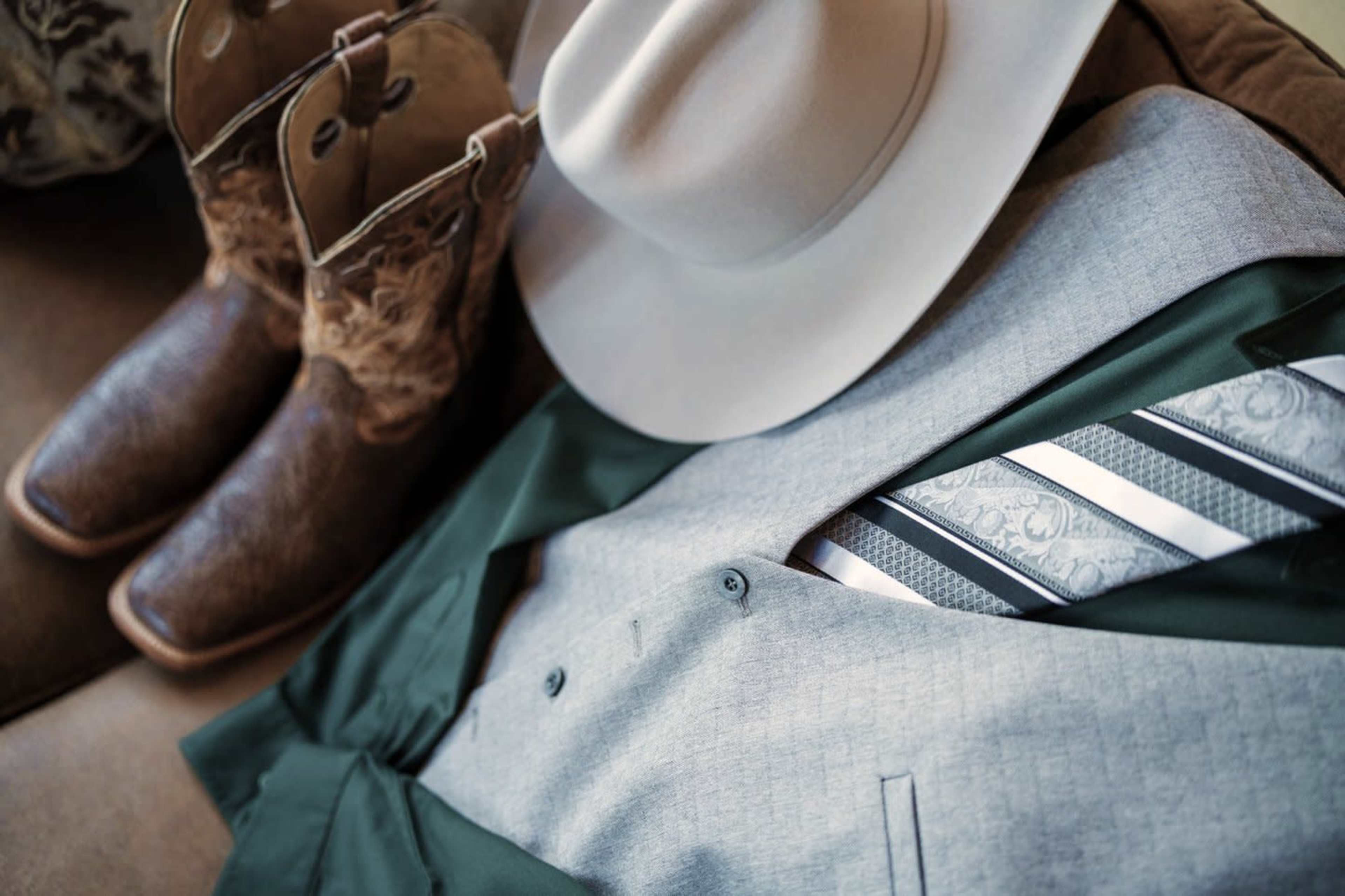 A gray suit with a patterned tie is arranged next to a cowboy hat and a pair of brown leather boots.