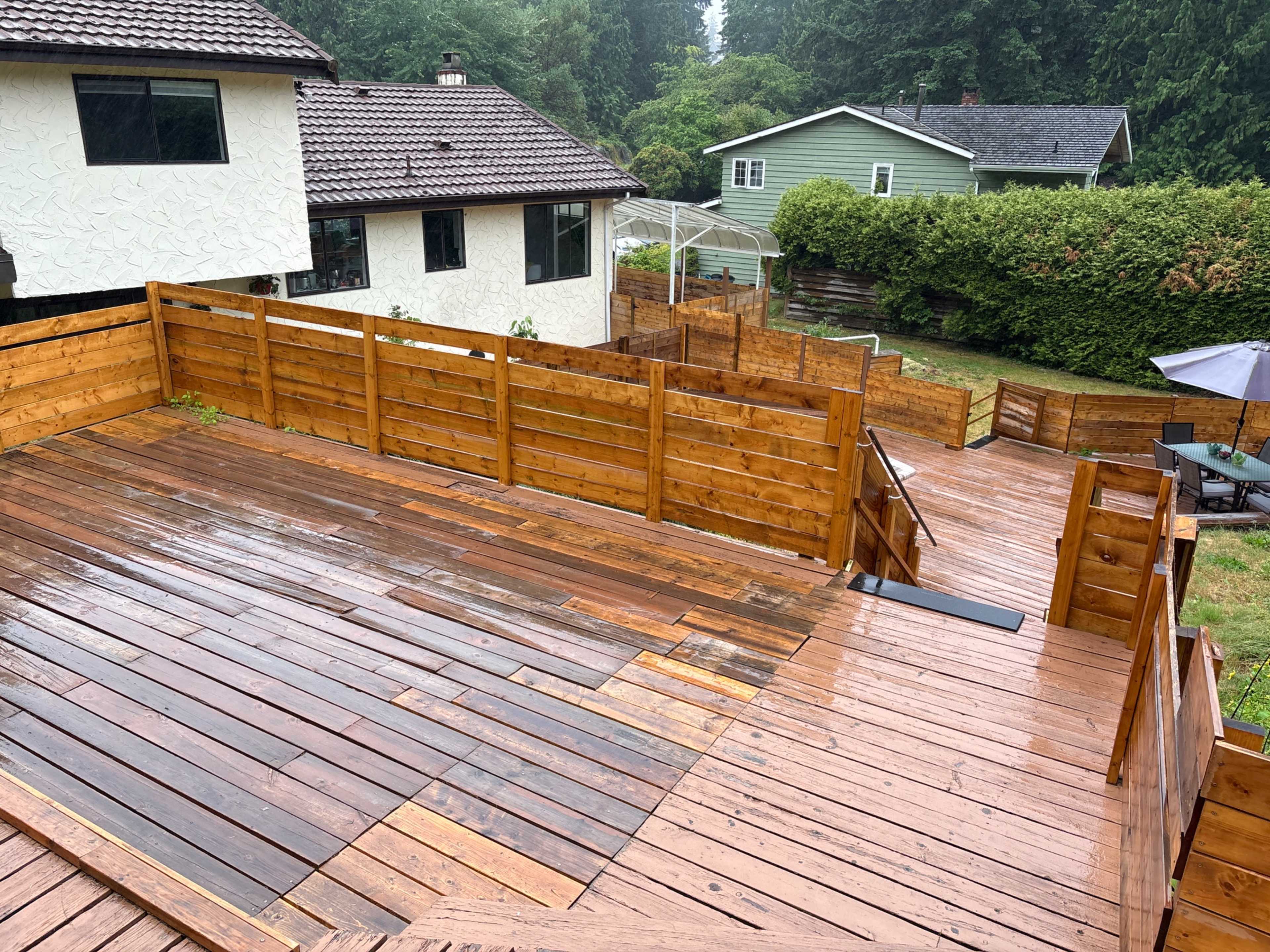 The image shows a wooden deck in a residential backyard, surrounded by houses and green foliage, with a wet surface indicating recent rain.