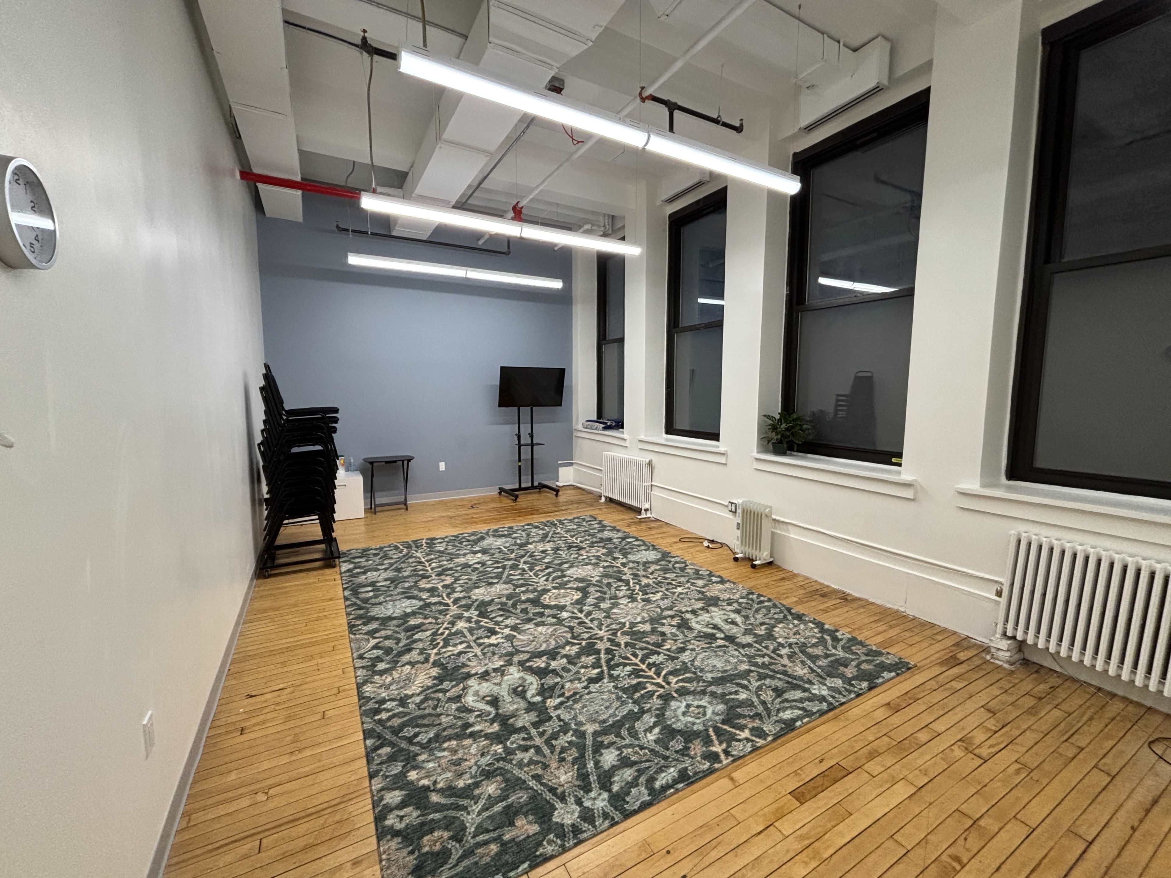 The image shows a sparsely furnished room with wooden floors, a large area rug, a television on a stand, and stacked chairs against one wall.