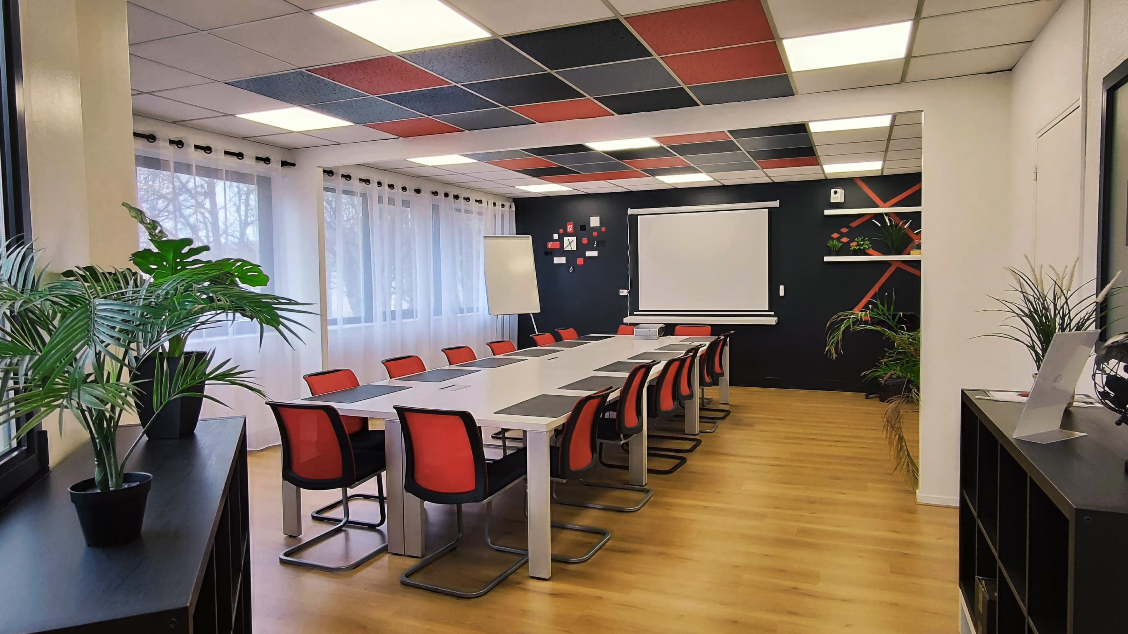 A modern conference room with a long table surrounded by red and gray chairs, a projector screen on the wall, and large windows letting in natural light.