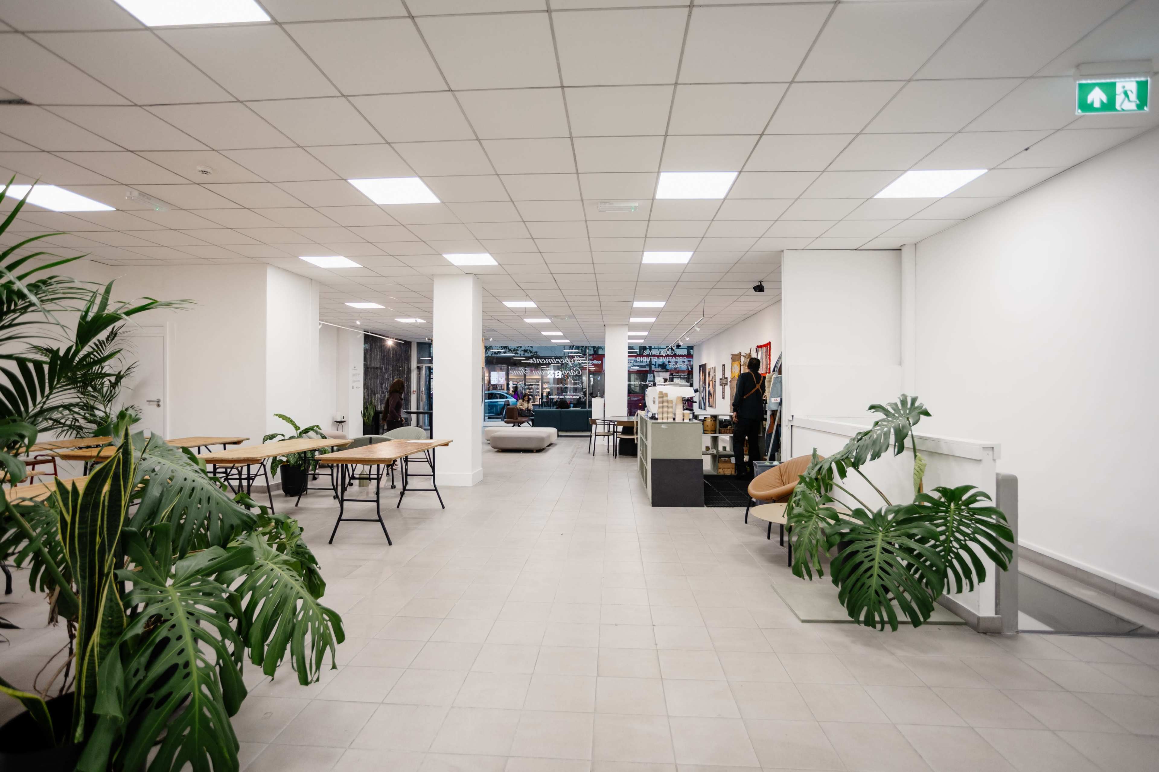 The image shows an open, well-lit interior space with several tables, plants, and minimalistic furniture.