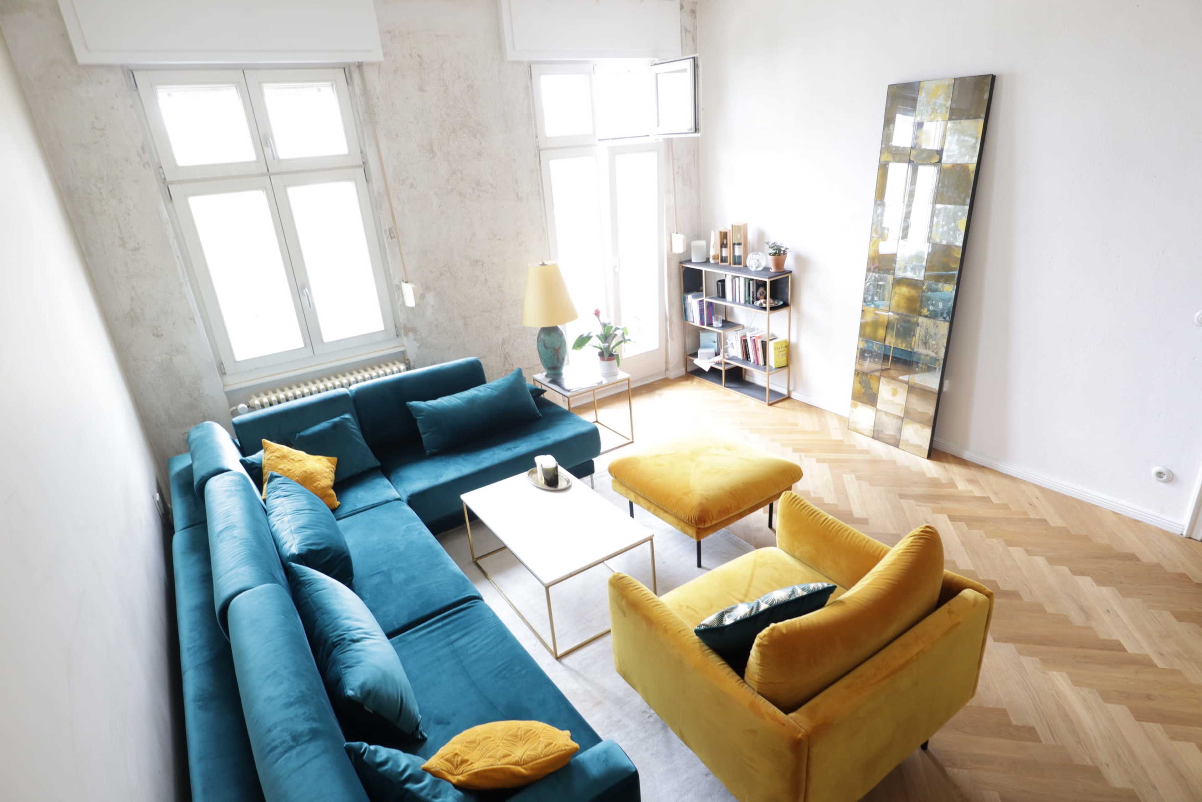 A modern living room with a blue sofa, a yellow ottoman, a coffee table, and a bookshelf against a light-colored wall with large windows.