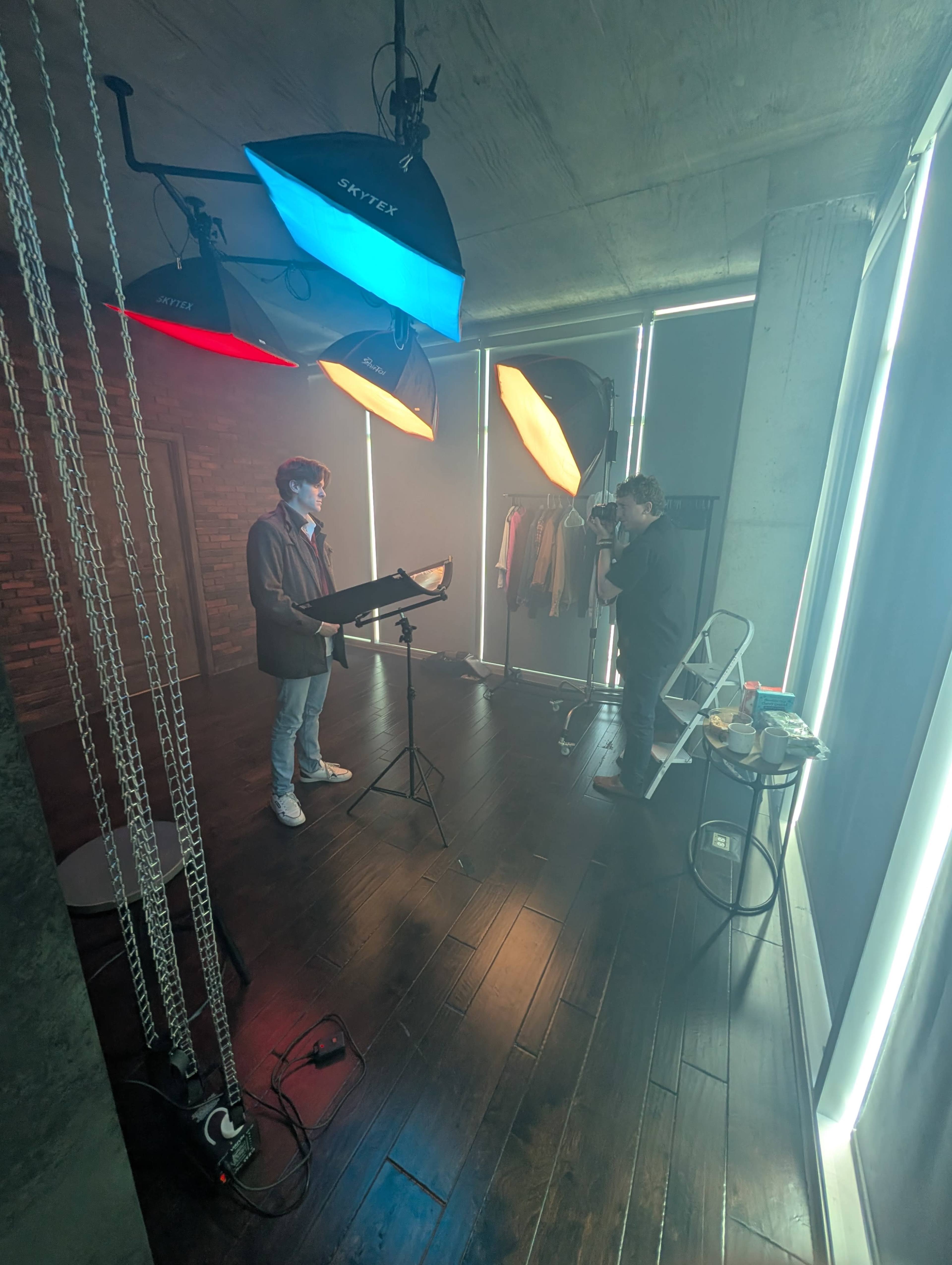 A person stands at a music stand under colored lights in a studio, while another person operates a camera nearby.