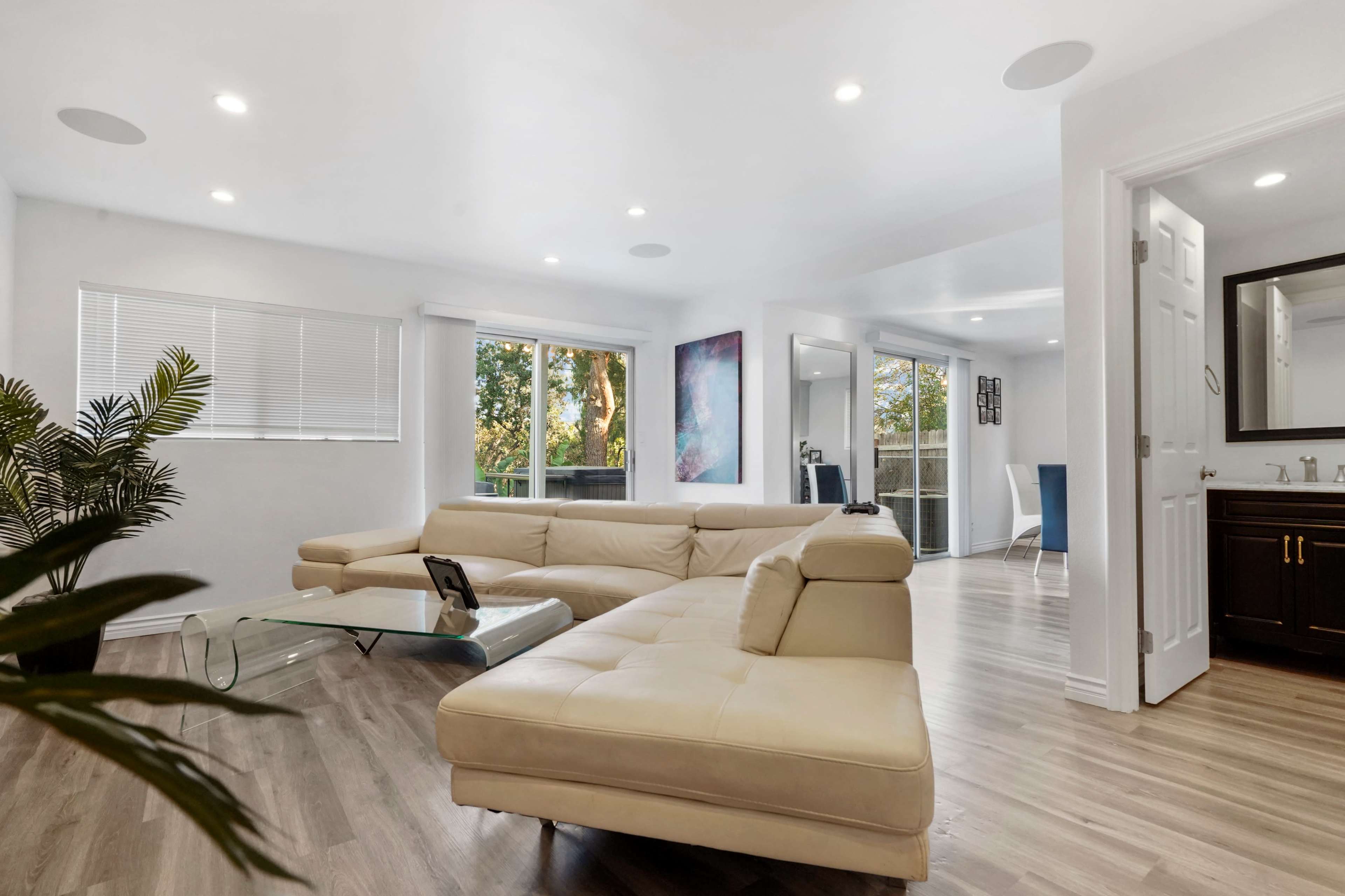 A modern living room with a light-colored sectional sofa, large windows allowing natural light, and an open layout leading to a dining area.