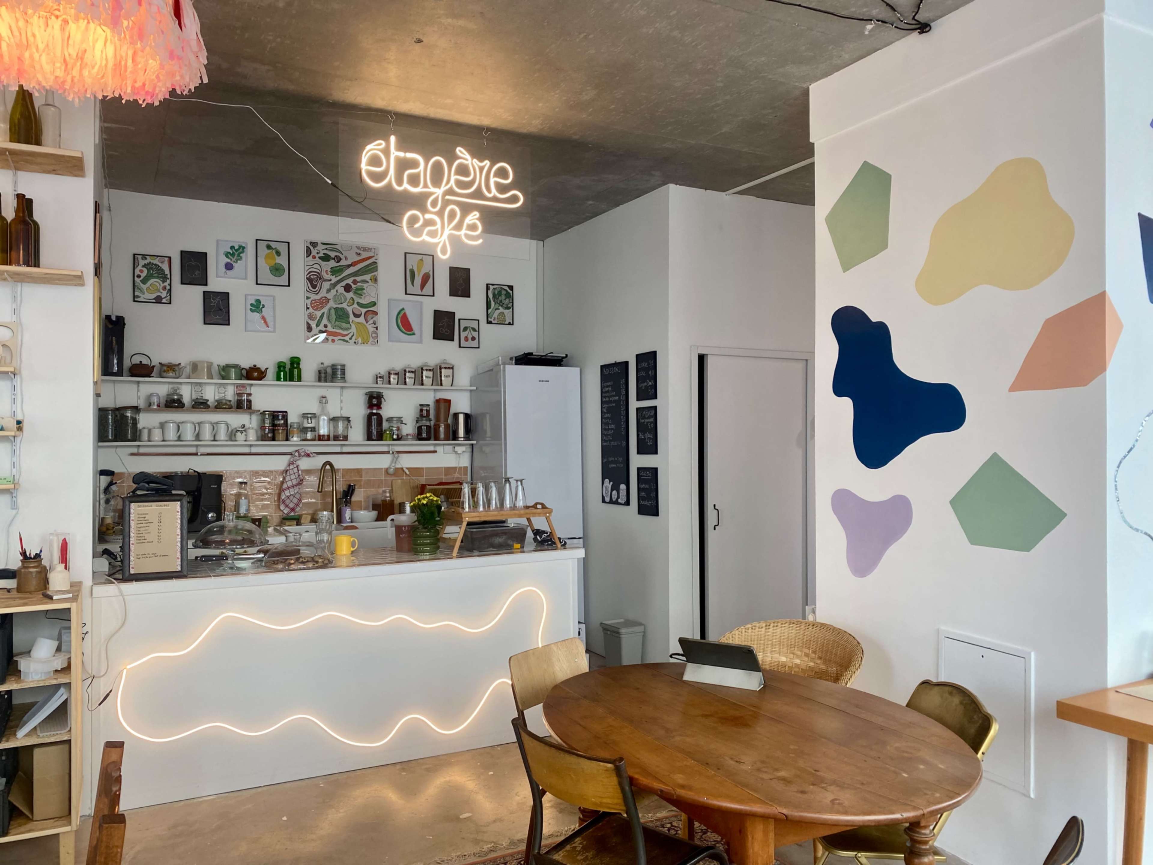 A cozy café interior featuring a counter with a neon sign, a round wooden table, and colorful abstract shapes on the wall.