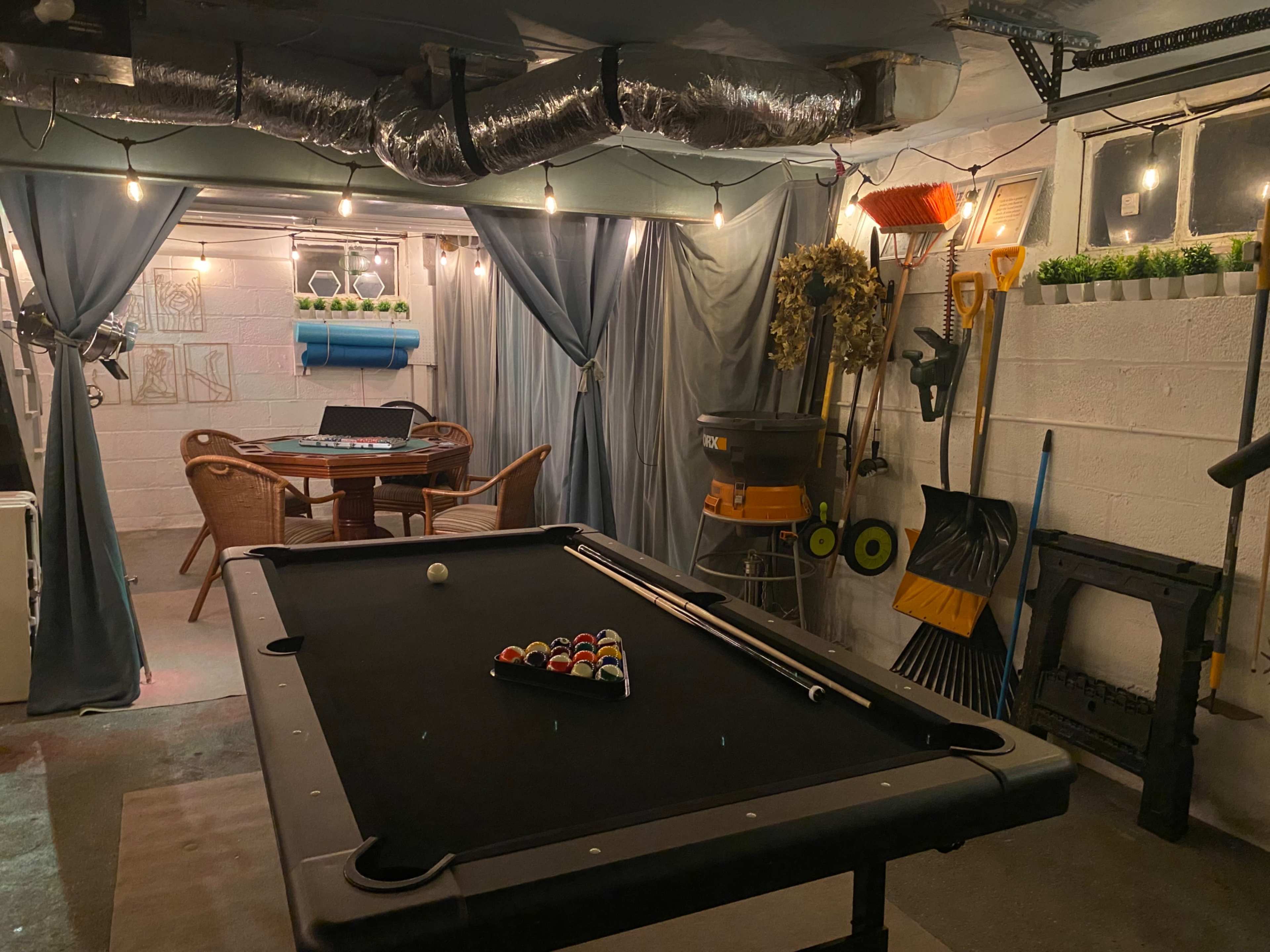 A dimly lit basement features a pool table with a triangle of balls, surrounded by chairs and tools hanging on the wall.