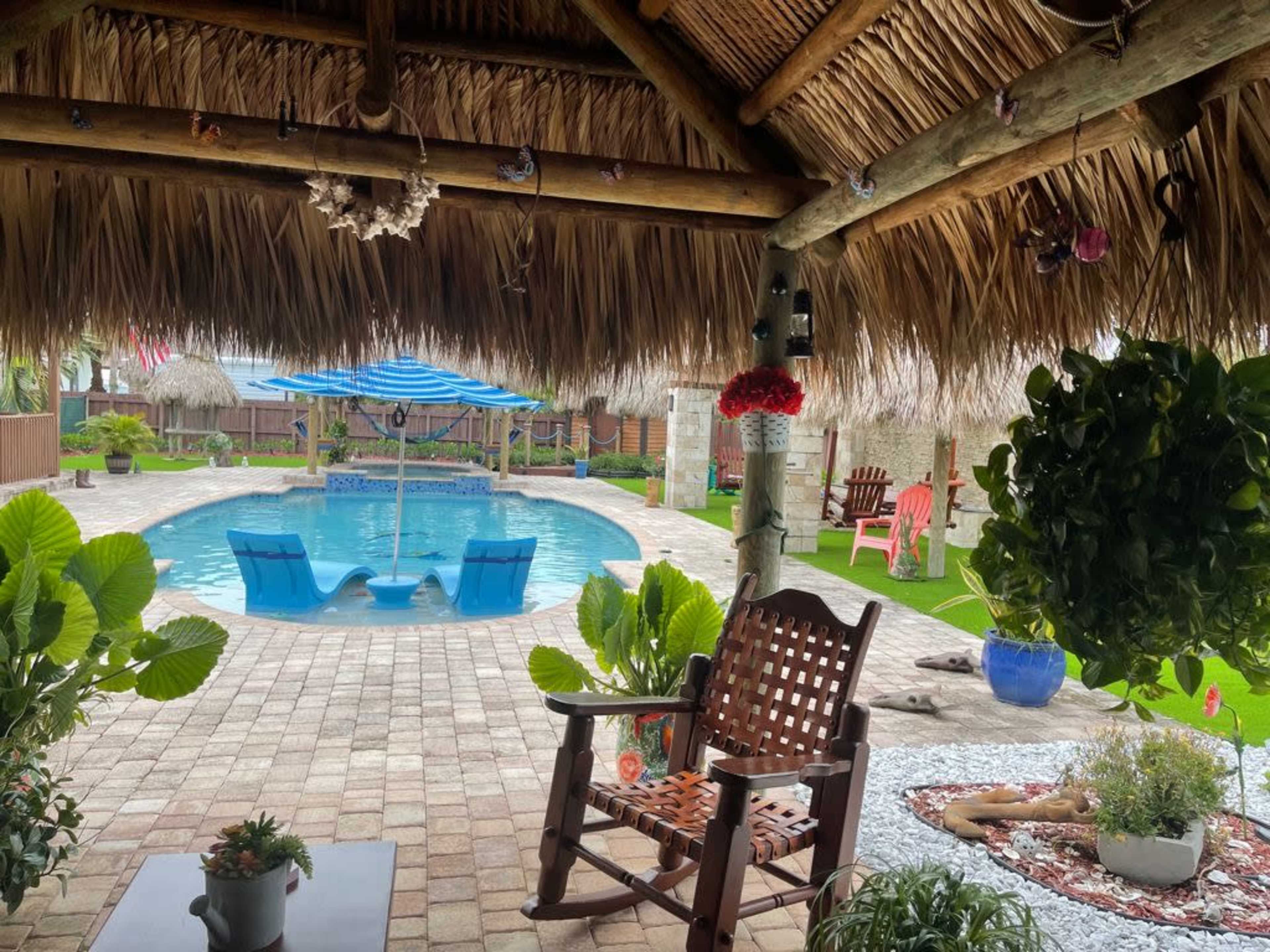 The image shows a backyard patio with a thatched roof, a swimming pool in the center, and lounge chairs beside it, surrounded by greenery and decorative elements.