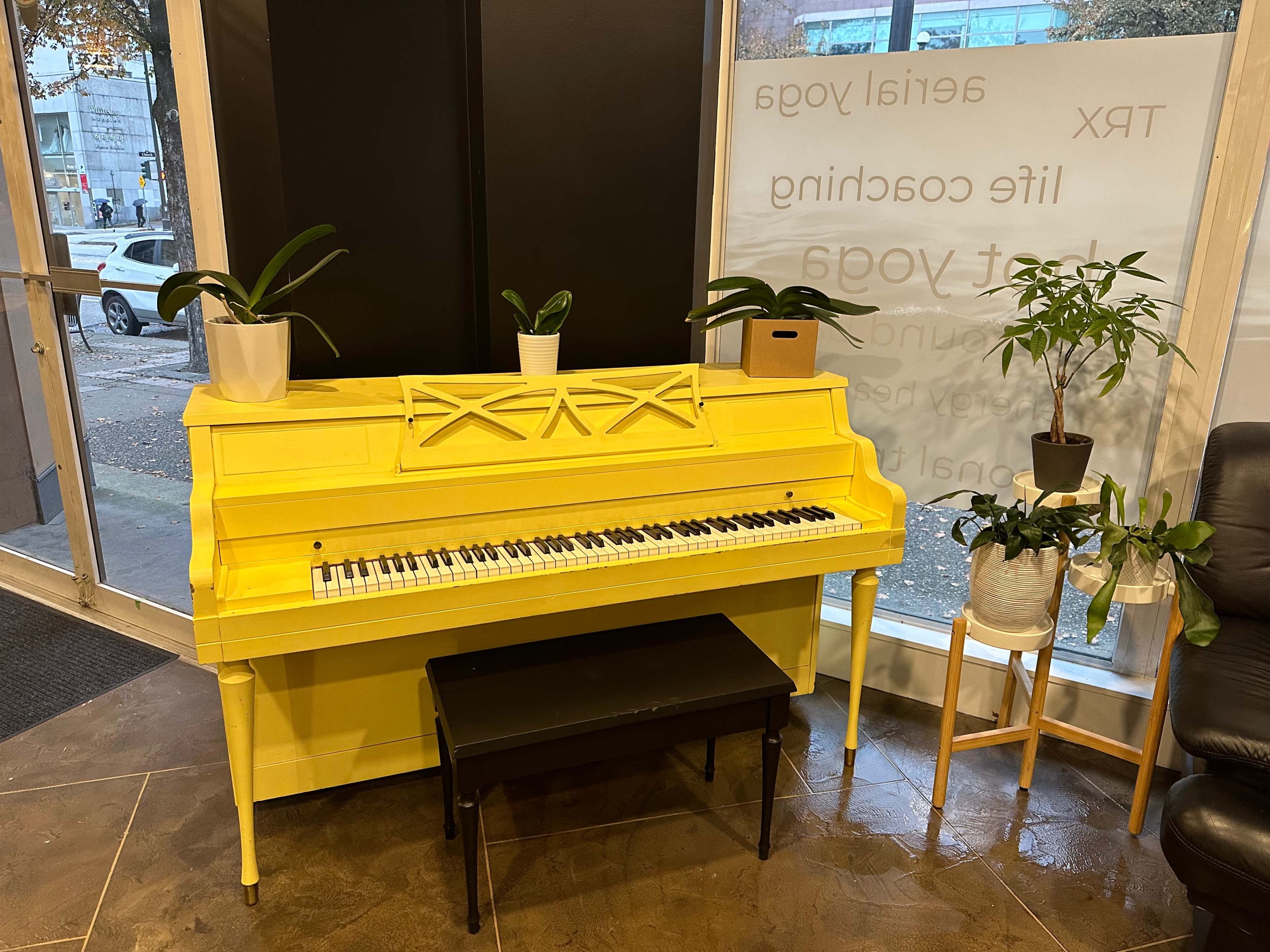 A yellow upright piano positioned next to two potted plants and a black bench in a well-lit interior space.