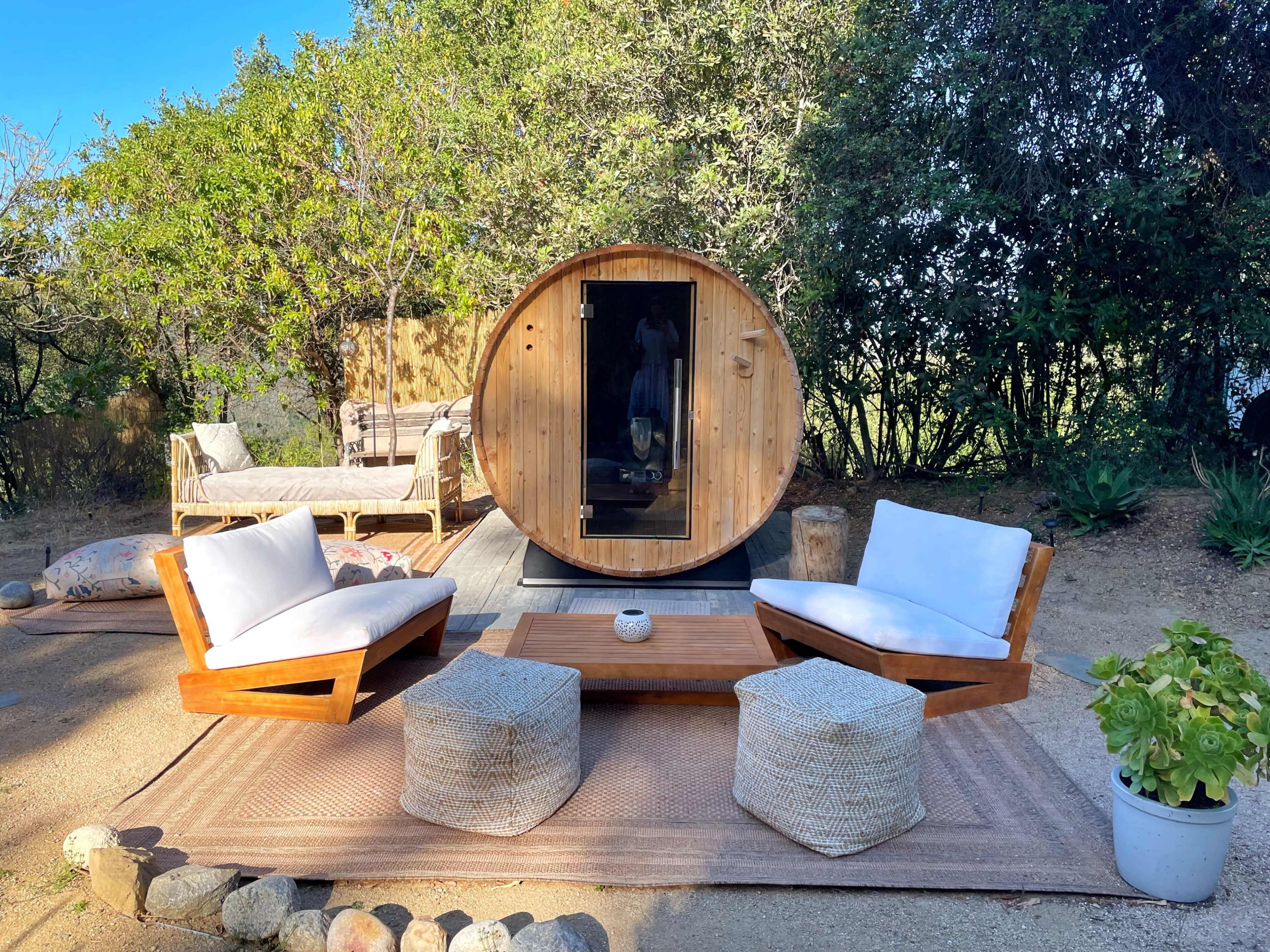 The scene features a wooden barrel sauna surrounded by outdoor seating, a coffee table, and lush greenery.
