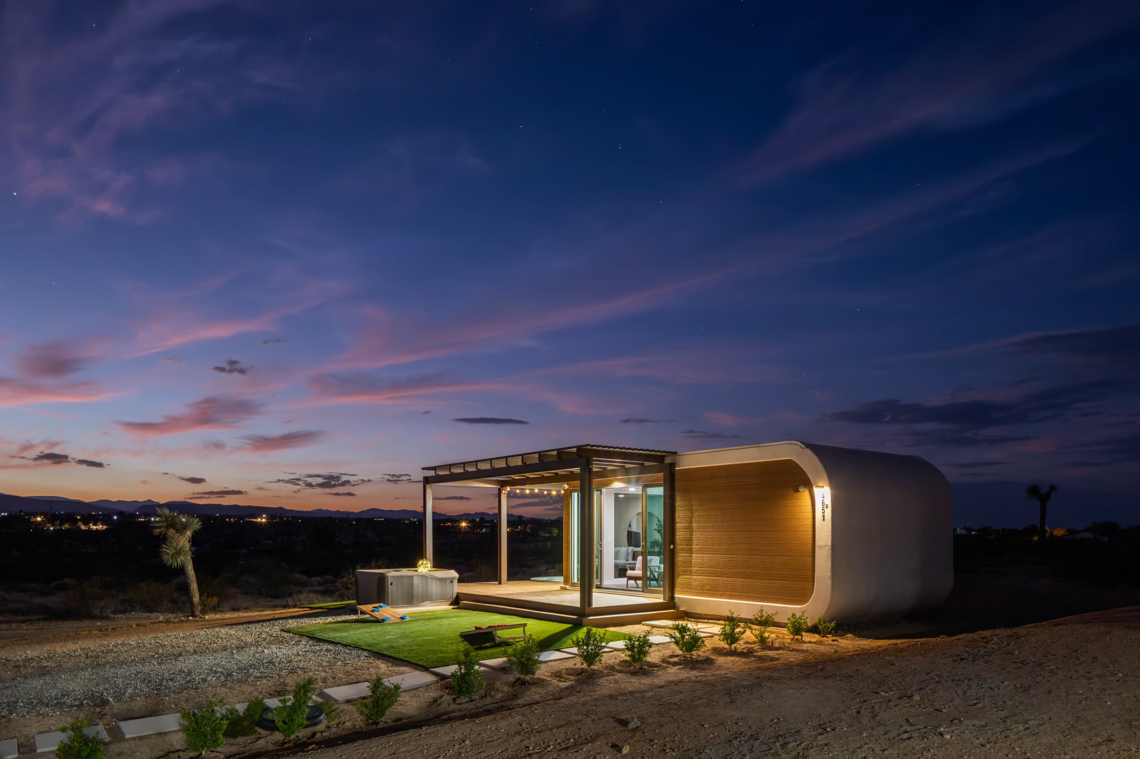 A modern, curved structure with large glass windows is set in a desert landscape at dusk, highlighted by soft exterior lighting.