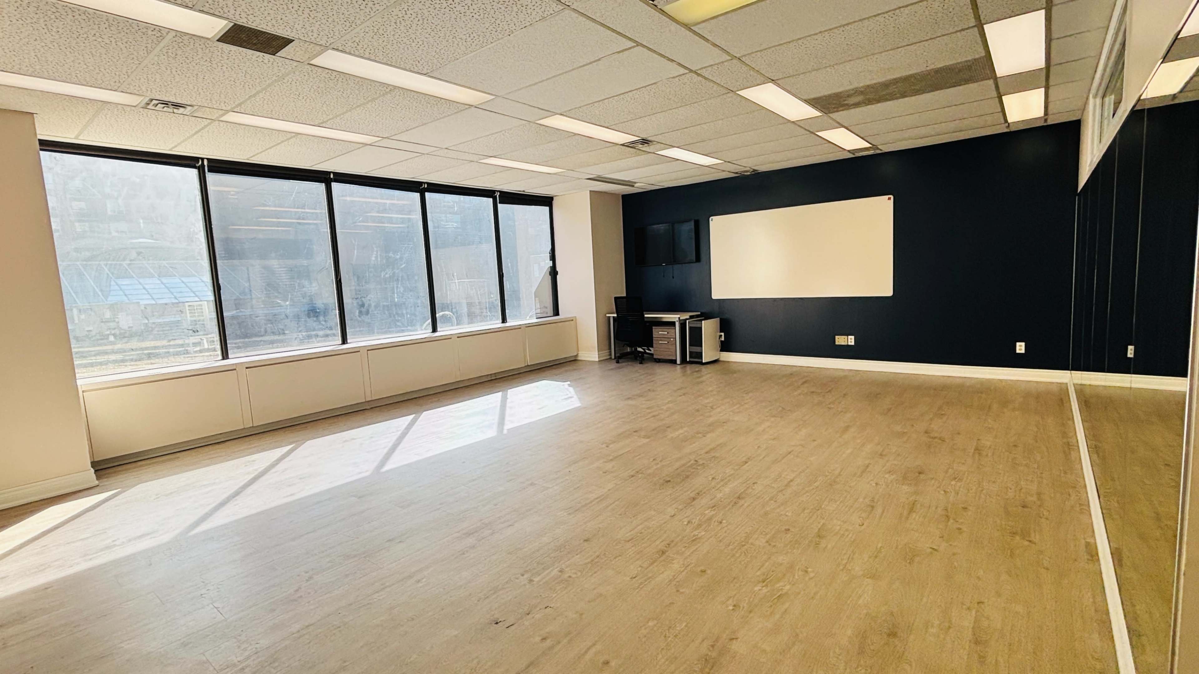 The image shows an empty room with large windows on one side, a dark feature wall, hardwood flooring, and a whiteboard mounted on the wall.