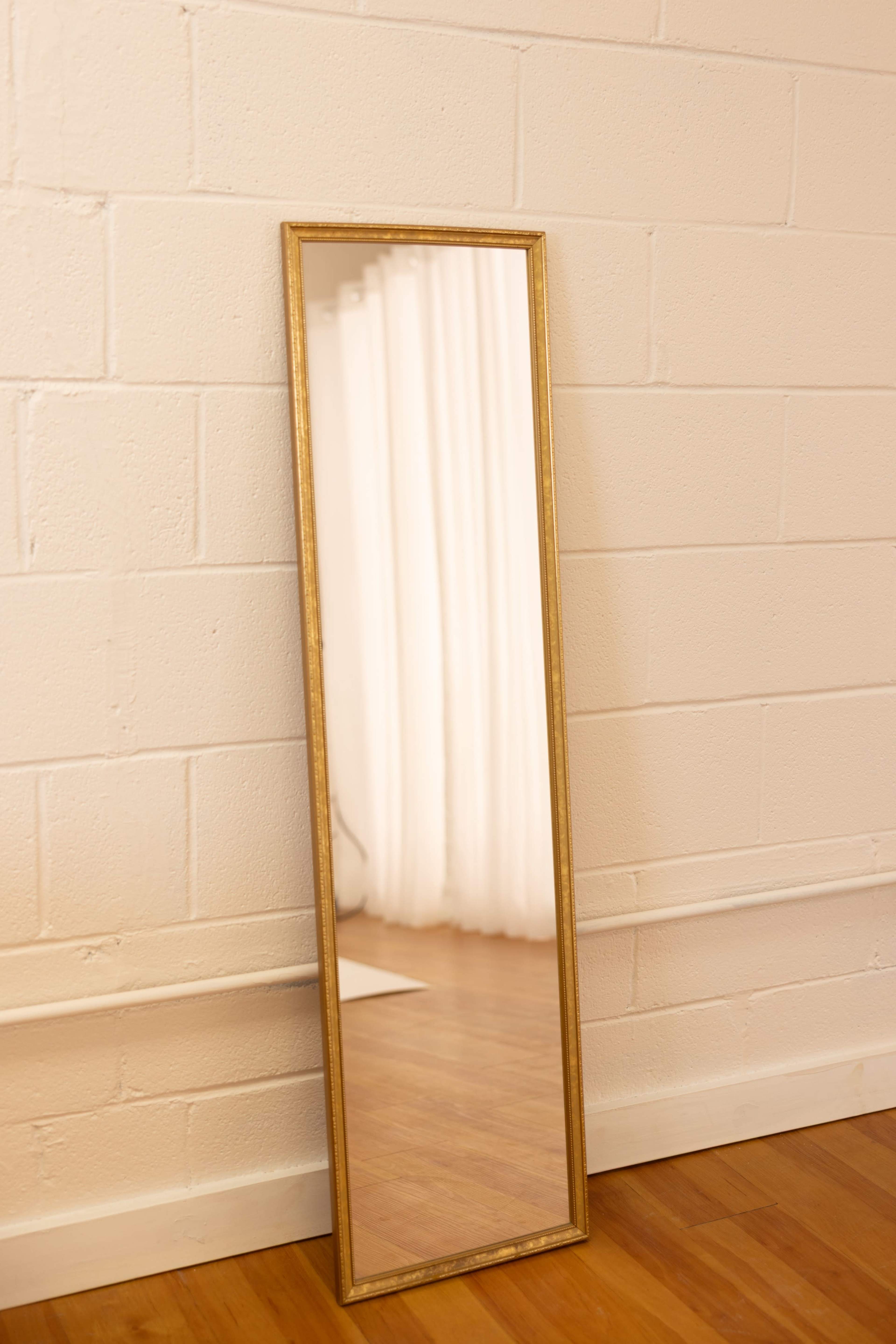 A tall, rectangular mirror in a gold frame leans against a white wall next to a wooden floor and partially visible curtains.