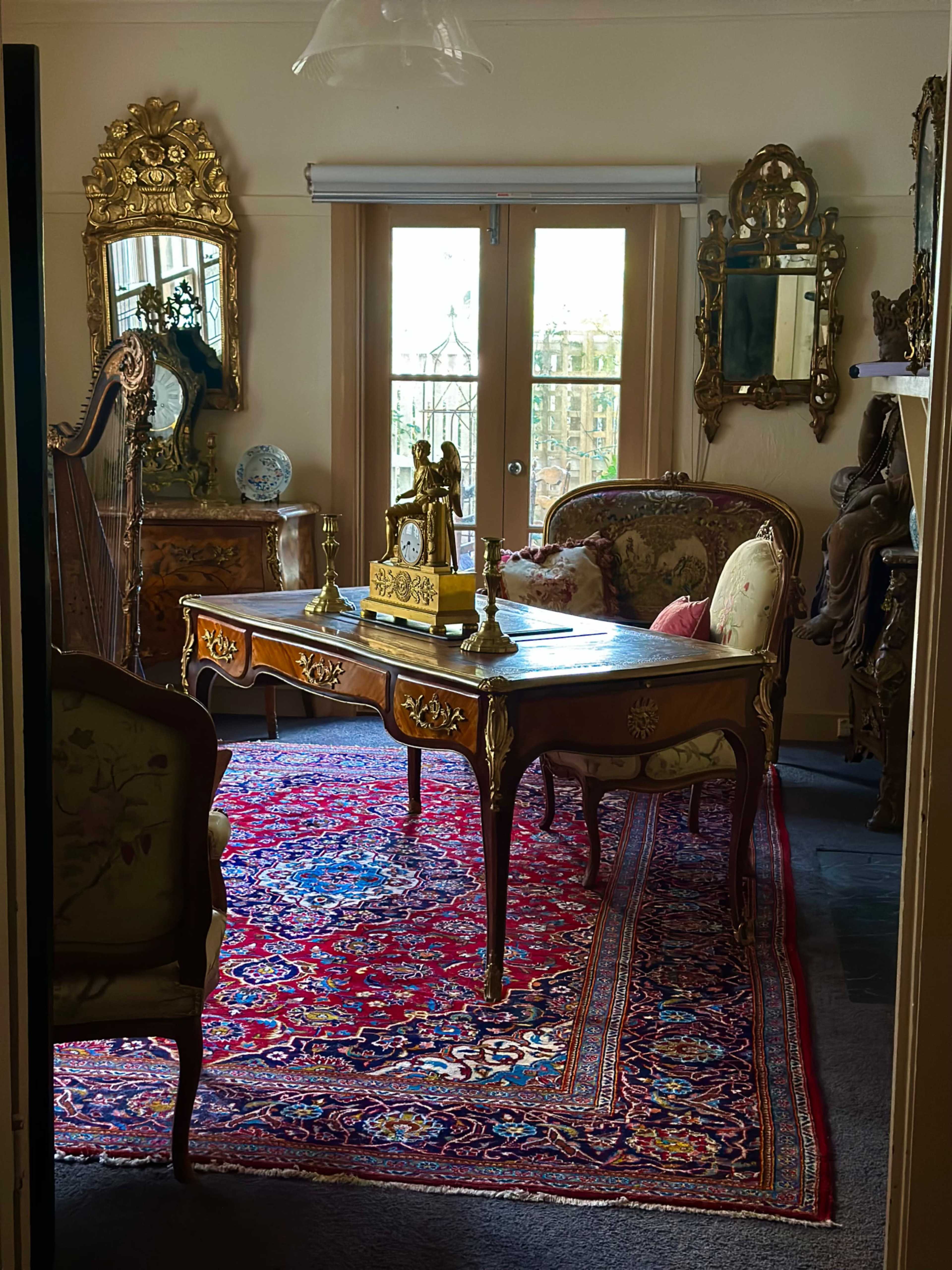 The image shows an ornate home office with a decorative wooden desk, a vintage clock, and elegantly styled chairs on a Persian rug, surrounded by mirrors and antique furnishings.