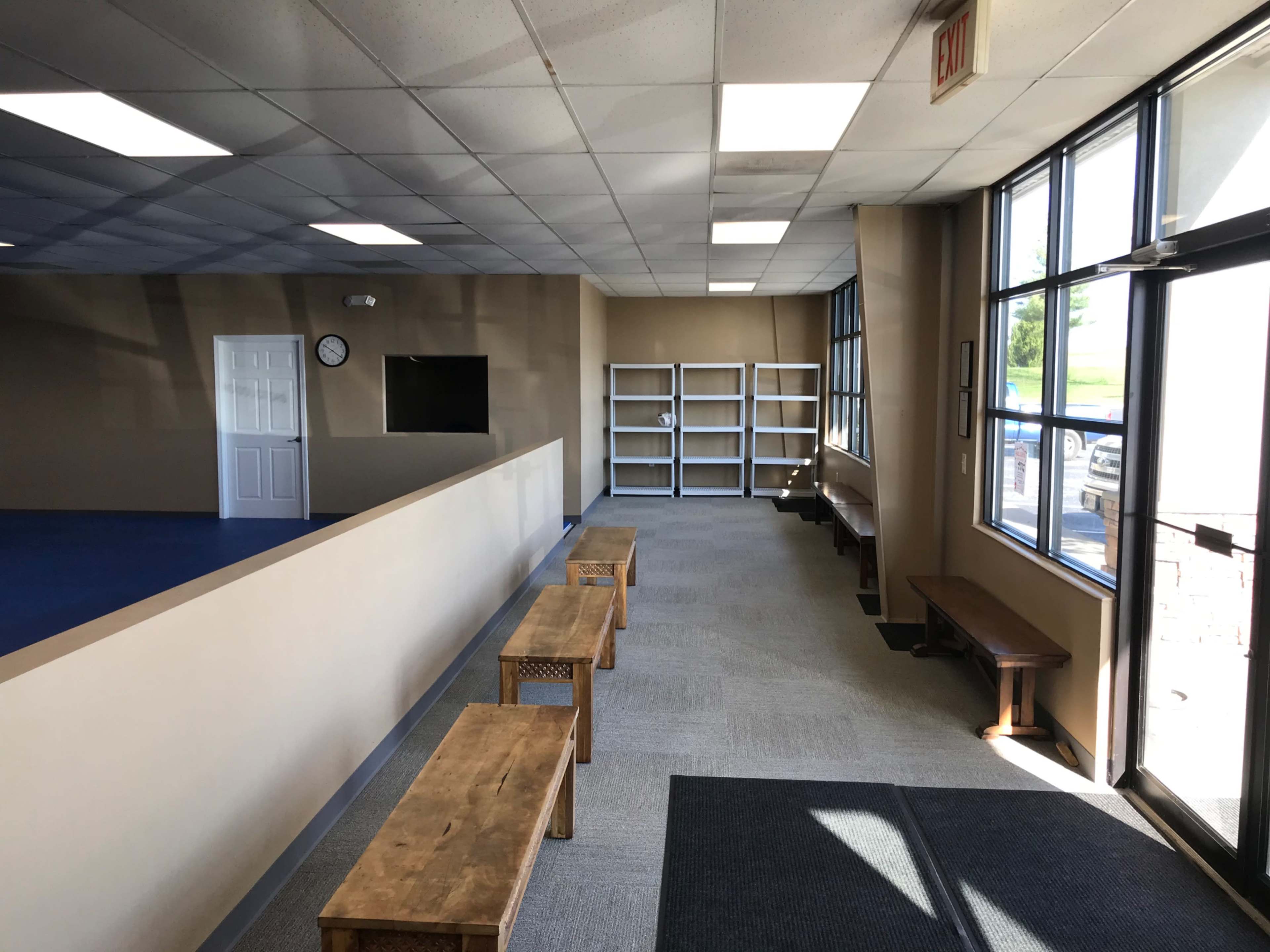 The image shows an interior space with a long, empty hallway, wooden benches along the sides, and shelves at the back against a beige wall.