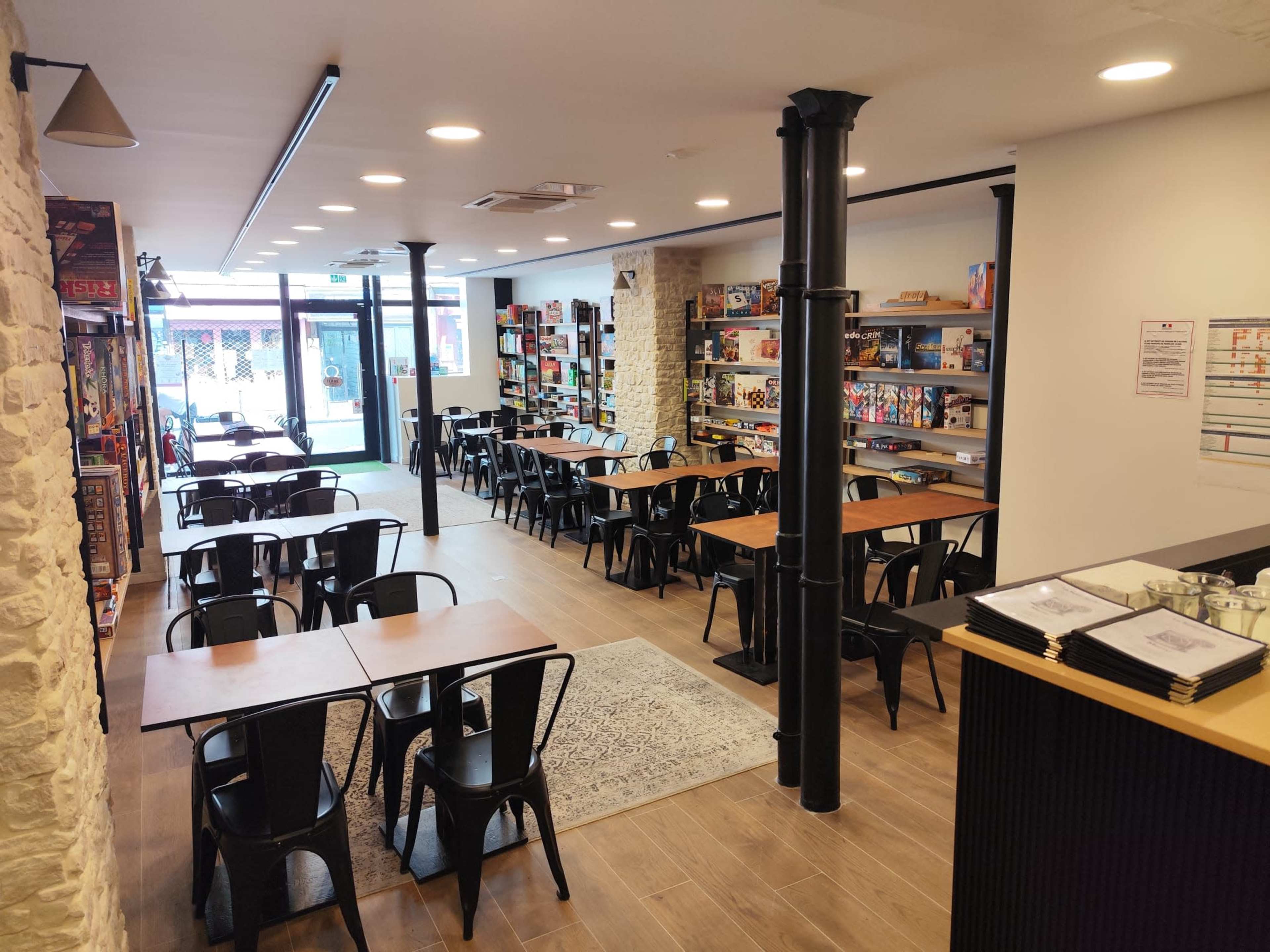 The image shows a spacious, brightly lit café with wooden tables and black chairs arranged neatly, along with shelves filled with board games along the walls.