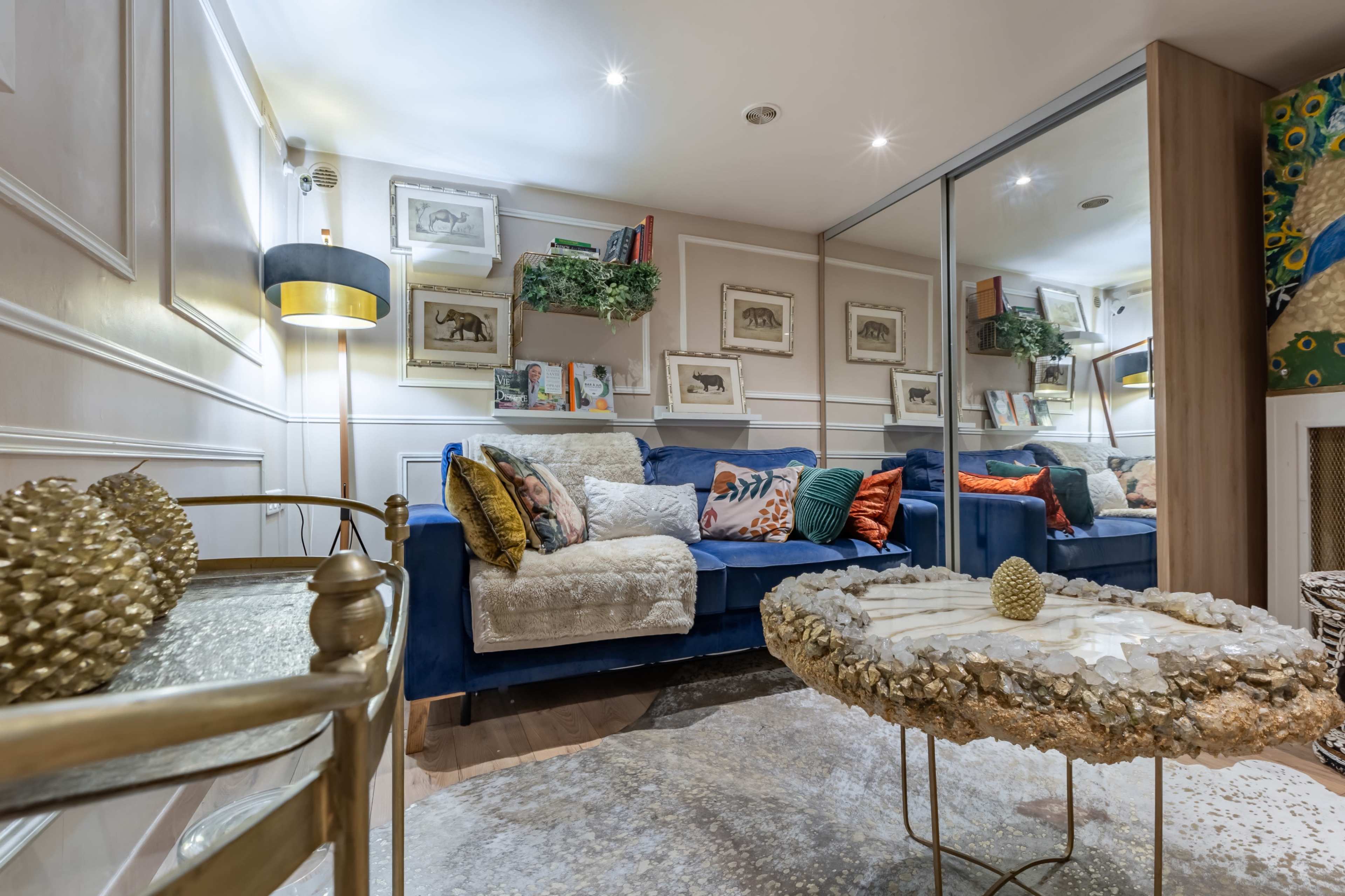 The image shows a cozy living room with a blue sofa adorned with colorful pillows, decorative shelves, and a unique coffee table set on a light rug.